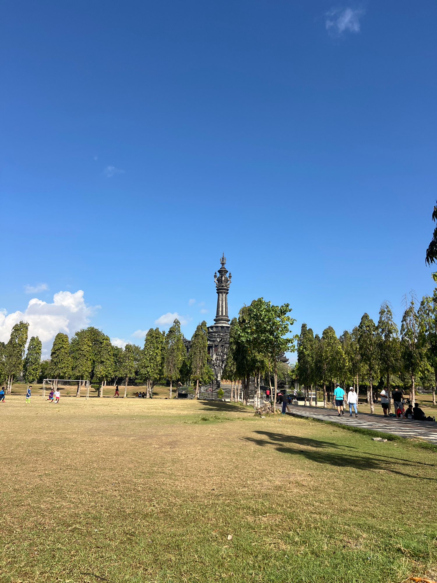 The image depicts the Bajra Sandhi Monument, a prominent landmark located in Denpasar, Bali, Indonesia. The monument, a tall, dark stone structure with intricate carvings and a spire-like top, stands centrally in the background, flanked by rows of lush green trees. In the foreground, a vast, dry grassy field stretches across the frame, dotted with a few patches of greener grass. On the left side of the field, a small group of children are engaged in a soccer game near a soccer goal. Further back, a paved walkway runs parallel to the trees, where several individuals are seen walking and some are sitting on the ground. The sky above is a clear, vibrant blue with a few wispy white clouds. The overall scene suggests a sunny day in a public park or recreational area.