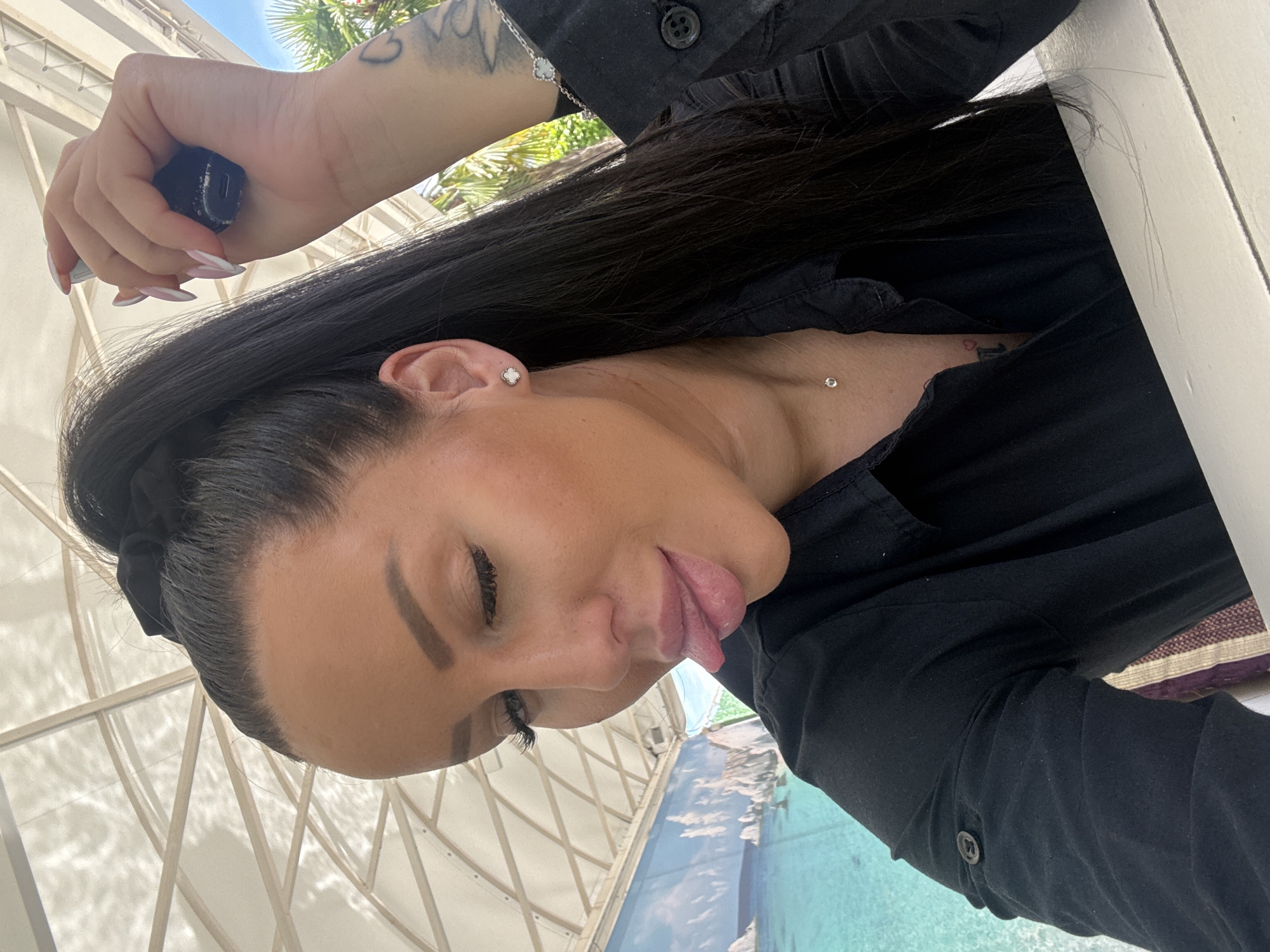 A woman with dark hair, fair skin, and manicured nails is holding a small, dark object in her left hand. She is wearing a black shirt and has a small diamond stud earring in her left ear, as well as a small diamond necklace, and a small tattoo on the center of her neck. Her tongue is sticking out. The background shows what appears to be an outdoor setting with a light-colored awning and greenery, as well as a body of water resembling a pool or pond with an underwater animal display, possibly in the city of Plovdiv, Bulgaria.