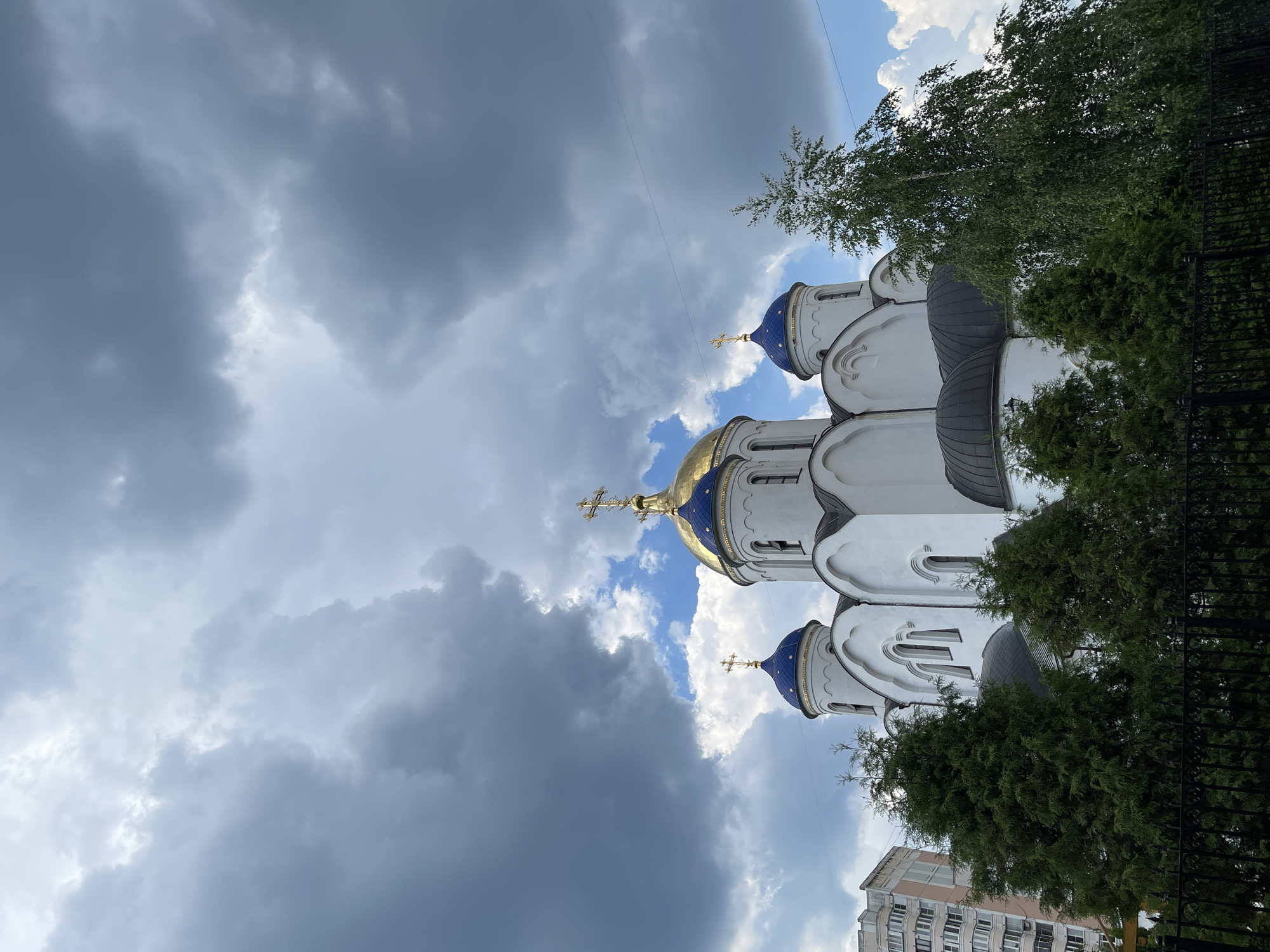The image shows a white Orthodox church with blue domes topped with gold crosses. The church is partially obscured by lush green trees and a black metal fence in the foreground. In the background, a tall, multi-story residential building is visible. The sky is dramatic, filled with dark, heavy clouds that suggest an impending storm, with patches of blue sky and white clouds visible between them. The overall impression is a religious structure set against a backdrop of both nature and urban development, under a moody sky.