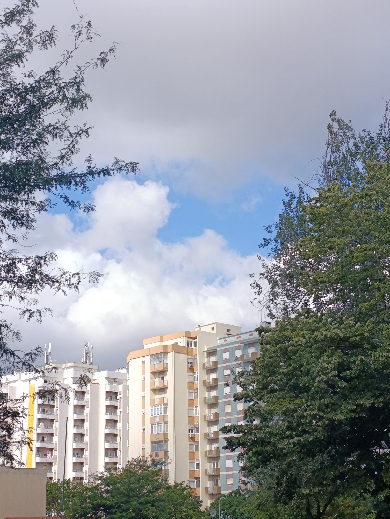 A low-angle, vertical, outdoor shot captures a cloudy sky with patches of blue, framing apartment buildings and trees in Setúbal, Portugal. The buildings, tall and light-colored, feature numerous balconies and windows. Trees with green foliage and thin branches are visible in the foreground and right side of the image, partially obscuring the buildings. The sky is predominantly overcast with large, white clouds, interspersed with areas of lighter blue sky. Several telecommunication antennas are mounted on the rooftops of some buildings. The overall scene suggests an urban environment under a mixed weather sky.