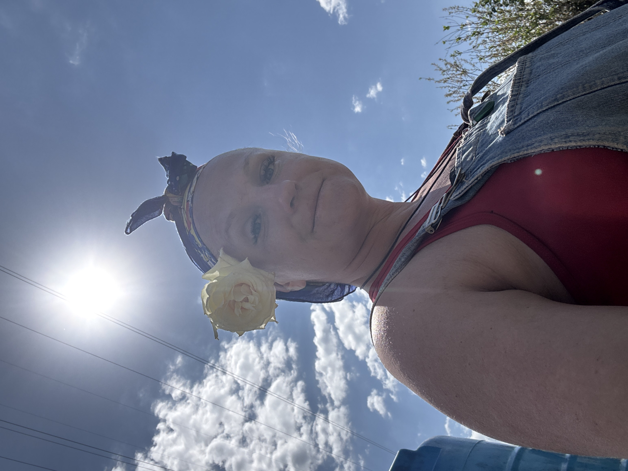 A woman, face tilted upwards, poses in front of a bright, sunny sky with scattered clouds. She wears a red top under denim overalls and a patterned headscarf. A light-yellow rose is positioned near her ear. Power lines stretch across the lower left portion of the sky. Based on visual clues, there is no information on the location being in the city of Chaikovsky, Russia.