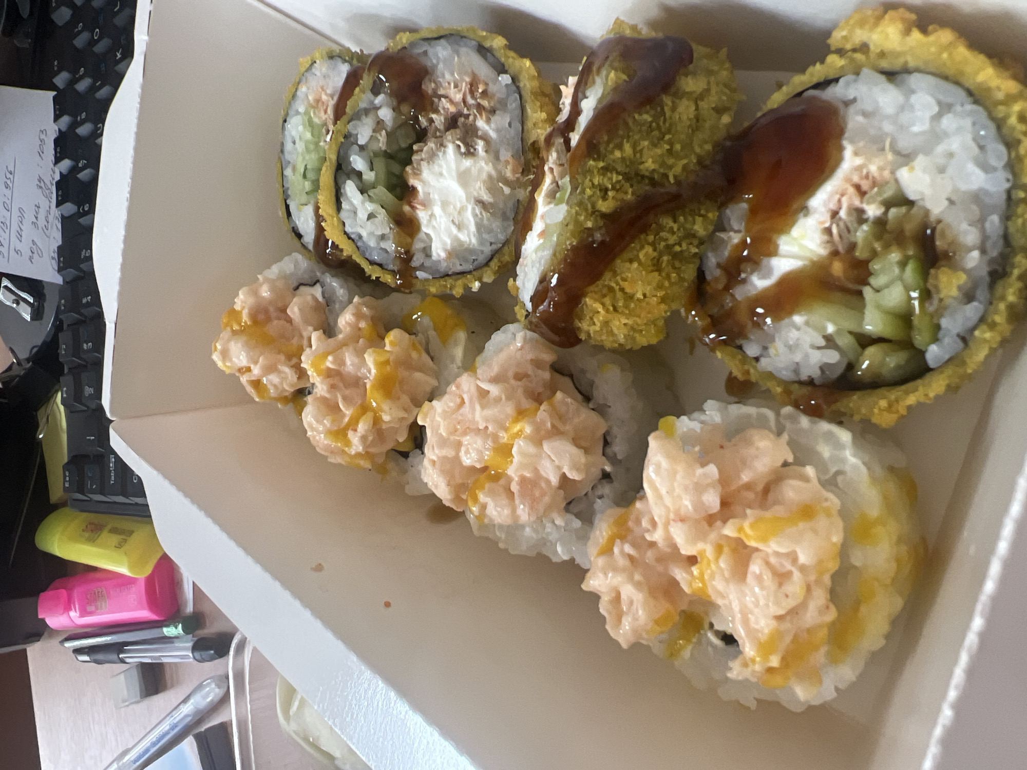 The image shows a white cardboard box containing several sushi rolls. The sushi rolls appear to be of two types: some are topped with a pink, creamy mixture and drizzled with a yellow sauce, while others are deep-fried and coated in a golden-brown breading, drizzled with a dark sauce. The box is placed on a desk with various office supplies visible in the background, including pens, highlighters, and a keyboard. A handwritten note is stuck to the keyboard. The lighting suggests an indoor setting, likely an office or home desk. There is no indication of the specific location within Volgograd, Russia.