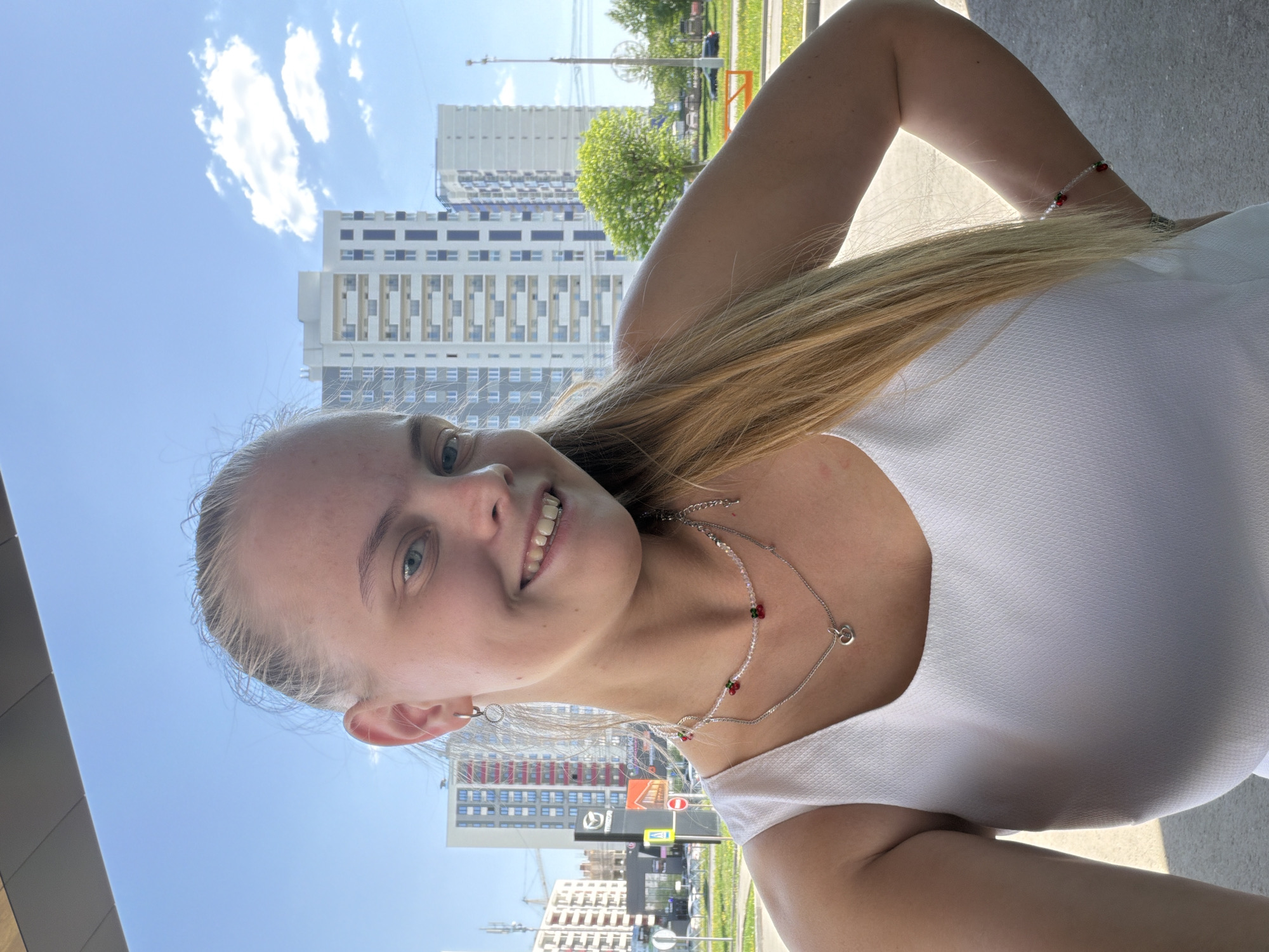A fair-skinned young woman with blonde hair is posing for a selfie in an urban environment. She is wearing a white top and two necklaces, one silver chain with a heart pendant and another beaded with red accents. She is smiling at the camera. In the background, there are high-rise apartment buildings and a blue sky with clouds. Street signs and traffic are visible, suggesting a city location.
Based on the style of architecture, signage, and vehicles, it is likely that this photograph was taken in Izhevsk, Russia. A Mazda dealership sign is visible in the background, which helps to identify the location as Izhevsk due to other corroborating details.