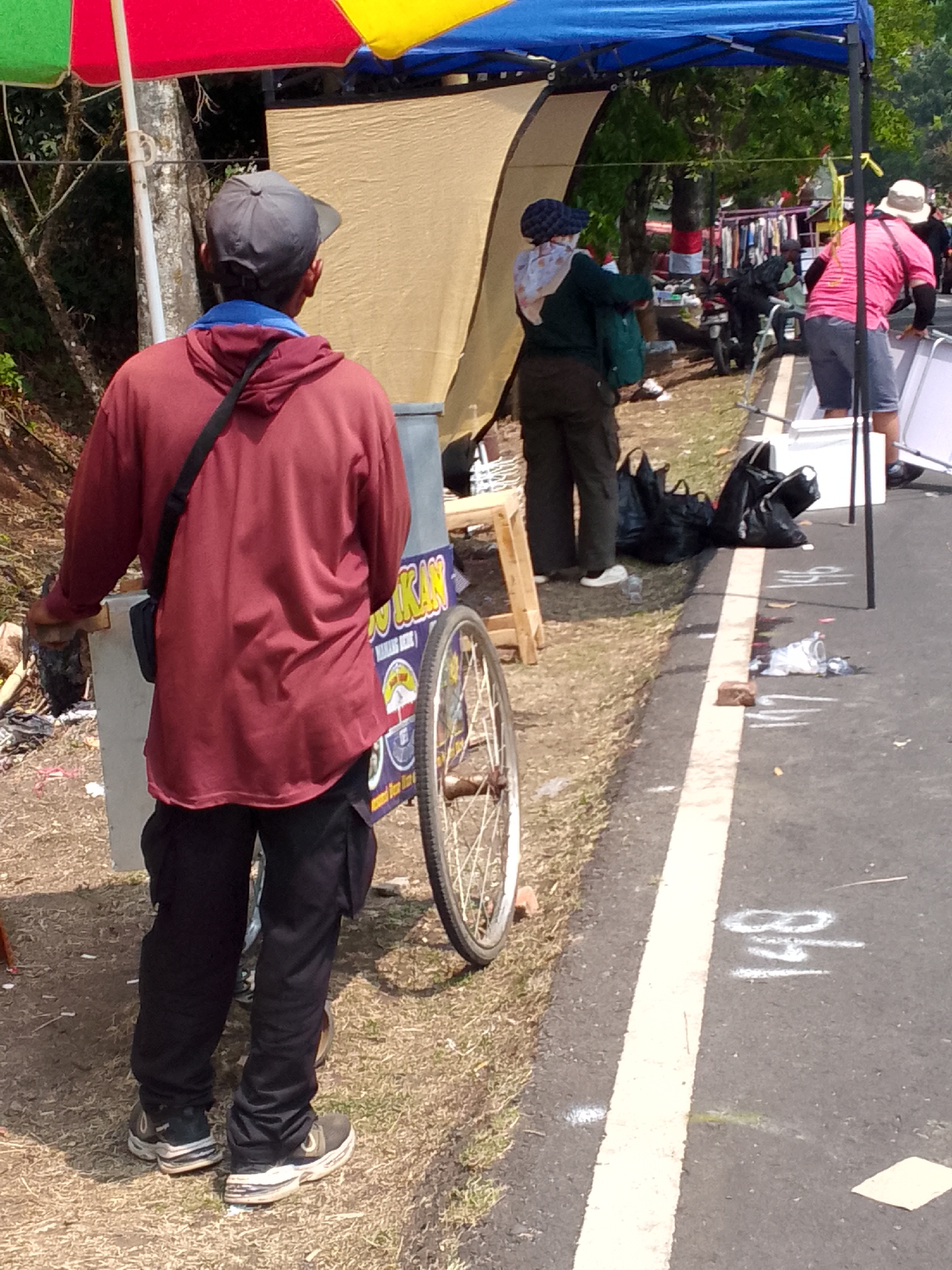 The image depicts an outdoor scene, likely in Mandalawangi, Indonesia, with a focus on street vendors and their carts. In the foreground, a man wearing a red hooded jacket and black pants stands with his back to the camera, near a cart with a wheel. He has a dark bag slung over his shoulder. The cart has colorful signage, partially obscured. Behind him, a woman in a green jacket and dark pants, wearing a headscarf, is also present. Further in the background, another person in a pink shirt and shorts is visible near a white tent or canopy. The environment appears to be a roadside, with a paved road marked with white lines and a grassy shoulder. Several black plastic bags are scattered on the ground. The overall scene suggests a market or a gathering of vendors.