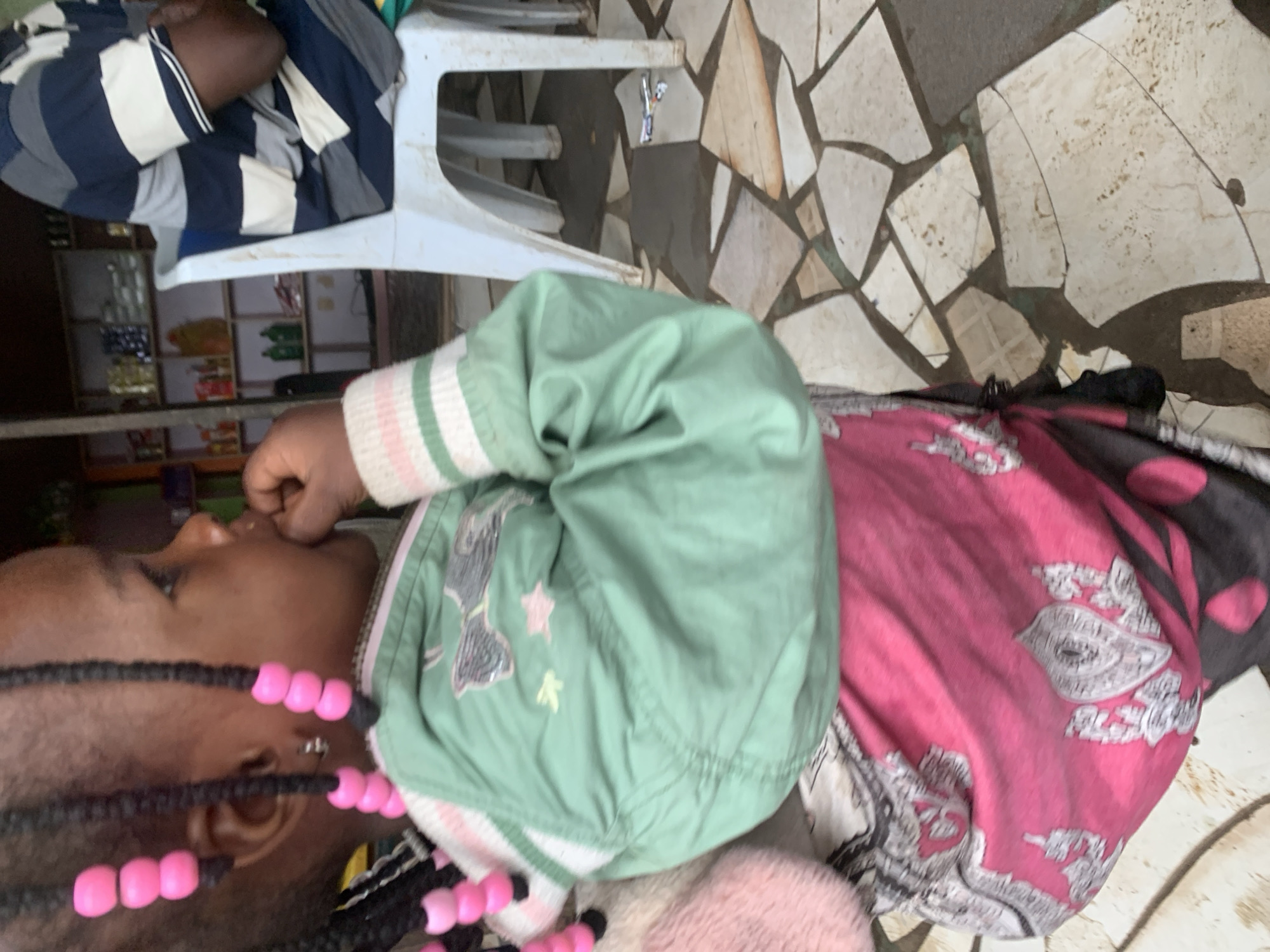A medium shot shows a young child with braided hair adorned with pink beads, wearing a green jacket with pink and white striped cuffs, and a pink and black patterned wrap. The child is seated, with their right hand near their mouth. To the right, a person in a blue and white striped shirt sits on a white plastic chair. In the background, shelves stocked with various items are visible, suggesting a shop or market stall. The ground is paved with irregularly shaped tiles. The scene is likely located in Shagamu, Nigeria, given the context.