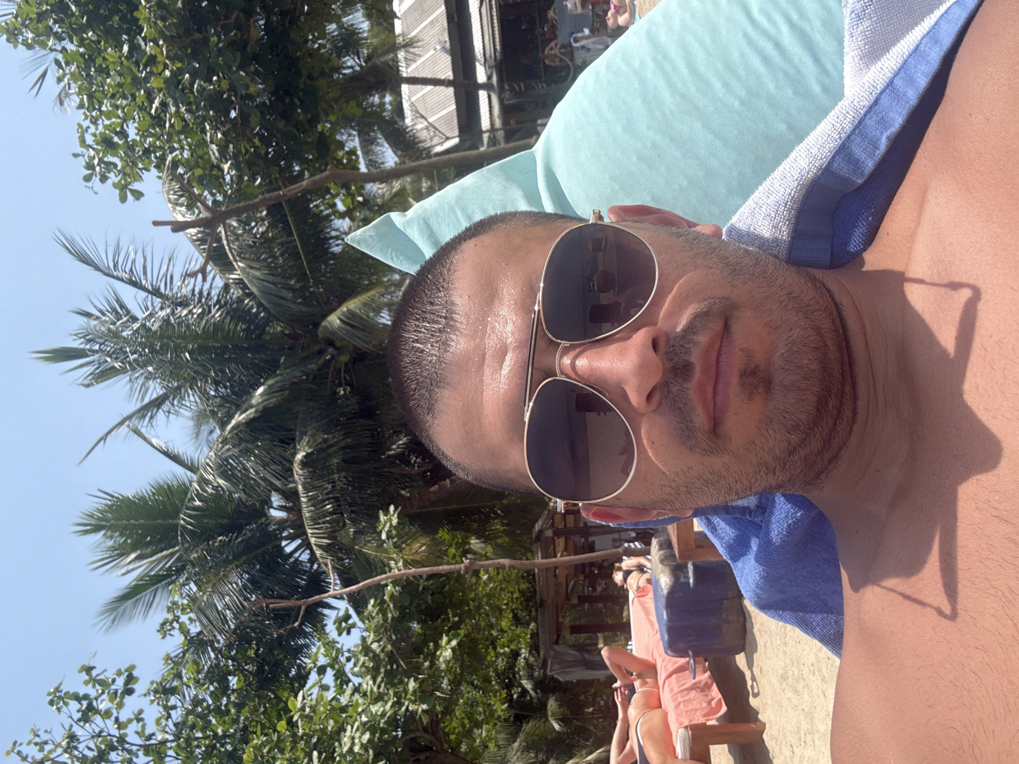 The image displays a selfie of a male individual taken on a developed section of a beach in Đà Nẵng, Vietnam, consistent with tourist areas like My Khe Beach.
**People:**
The main subject is a male, visible from the mid-chest upwards, positioned centrally. He has short dark hair, a stubble beard, and a tanned complexion. He is wearing gold-framed aviator sunglasses that reflect the surrounding environment. He is shirtless. In the background, on the sandy beach, at least two other individuals are visible. One person is partially obscured on the far left, lying face-down on a red-orange beach lounger, wearing dark swimwear. Another person is partially visible on the right, also lying on a red-orange beach lounger, wearing a pink or red swimsuit. Additional blurred figures are discernible further in the background near a building.
**Subjects/Objects:**
The main subject leans against a large, light blue beanbag pillow. A blue and white striped towel is draped over his left shoulder and chest. A blue cylindrical object, possibly a cooler or stand, is located next to a beach lounger in the mid-left background. Several red-orange beach loungers are scattered across the sandy area.
**Actions/Interactions:**
The main subject is actively taking a selfie. The other visible individuals are engaged in leisure activities, relaxing on beach loungers.
**Environment:**
The scene is set on a sandy beach. The sky is clear and bright blue, indicating sunny weather. Lush green tropical foliage, including prominent palm trees and other deciduous trees, dominates the upper and left background. In the mid-right background, a rustic beachside structure with a dark corrugated metal roof and wooden supports is visible, suggesting a beach bar or restaurant. The strong sunlight casts shadows, including one from a thin object on the main subject's chest and subtle shadows beneath trees and objects.