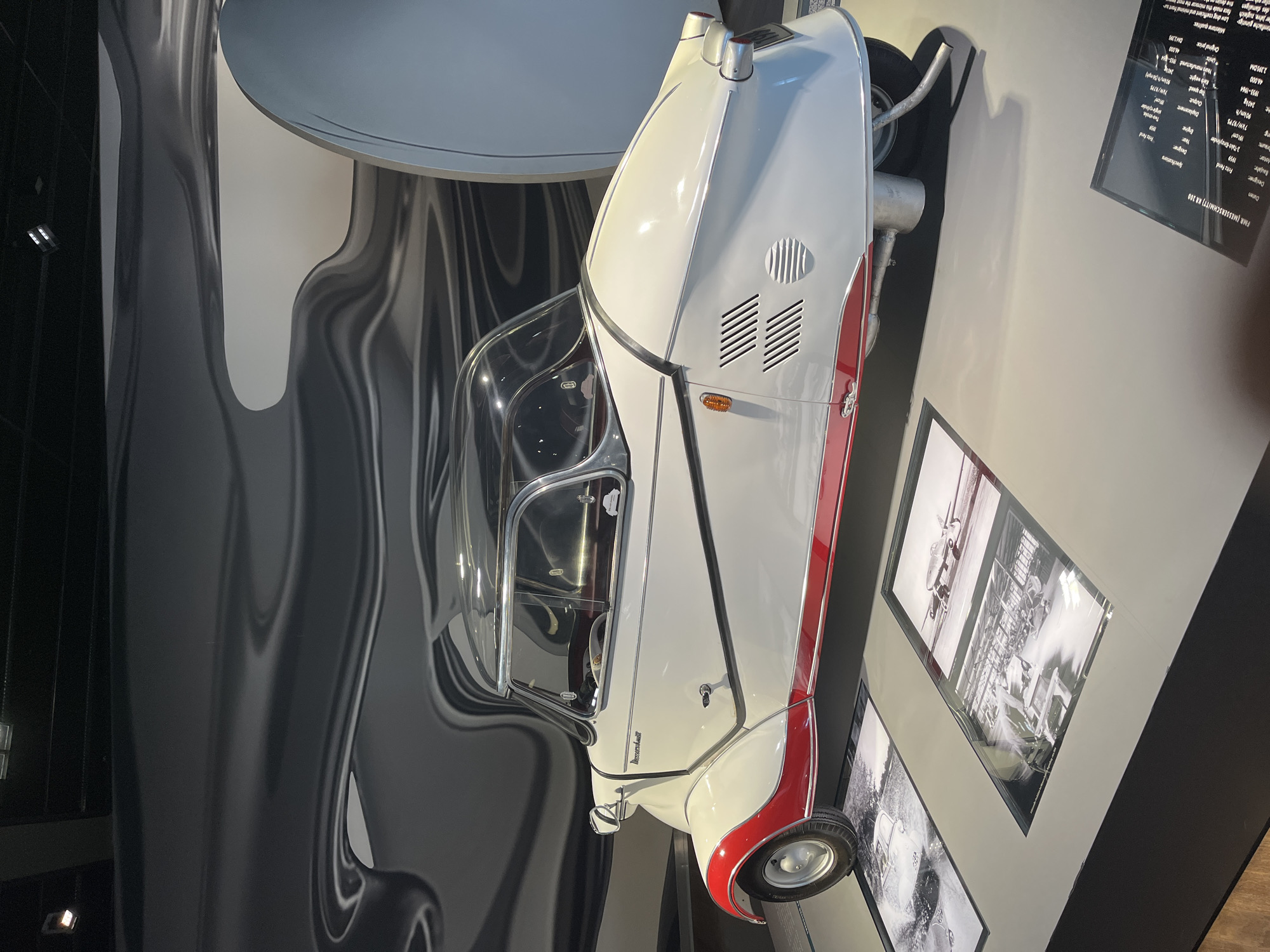 This is a medium-shot, eye-level, indoor photograph of a Messerschmitt KR200 three-wheeled microcar, which is a German vehicle. The microcar is white with red accents on the fenders and lower body panels, and has a clear canopy top. It is displayed on a grey platform, with several framed historical photographs displayed on the floor in front of it. The background features a large, abstract, metallic wall with swirling patterns. To the right of the microcar, a portion of a large, rounded, pale blue or grey structure is visible, possibly part of the museum's architecture. The overall scene suggests a museum or exhibition setting. The text on the display sign below the vehicle is partially visible and includes information about the vehicle's specifications and history, and the location is the Autostadt in Wolfsburg, Germany. There are no people visible in the image.