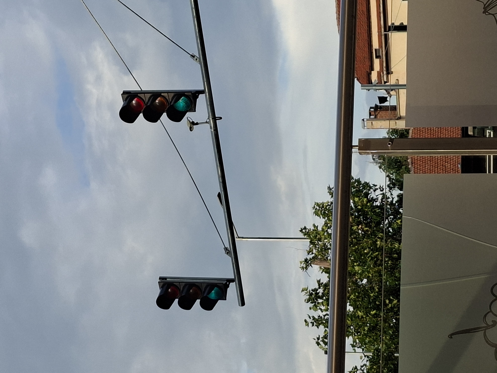 The image, correctly oriented, displays an urban street scene dominated by overhead infrastructure and architectural elements under a partly cloudy sky. Two sets of traffic lights are suspended from wires spanning across the frame.
The upper traffic light fixture shows its red and amber (yellow/orange) signals illuminated, while the green light appears off. The lower traffic light fixture, positioned further away, displays an illuminated green signal, indicating "go" for traffic. The traffic lights are black with visors over each lamp.
On the right side of the image, the upper stories of several buildings are visible. These buildings feature traditional terracotta-tiled roofs and light-colored facades, with some windows discernible. Green tree foliage is scattered throughout the right side, suggesting presence of greenery alongside the urban structures. A prominent metallic railing or architectural support structure runs vertically along the far right edge of the frame, alongside a reflective or translucent panel, possibly part of a building, balcony, or commercial awning.
The sky is a bright blue with wispy white clouds, indicating daytime conditions. There are no people or vehicles visible, focusing the scene on the urban landscape and its signaling systems. The setting appears to be a street or intersection within a city, possibly Plovdiv, Bulgaria, as indicated by the context. No visible text is present in the image.