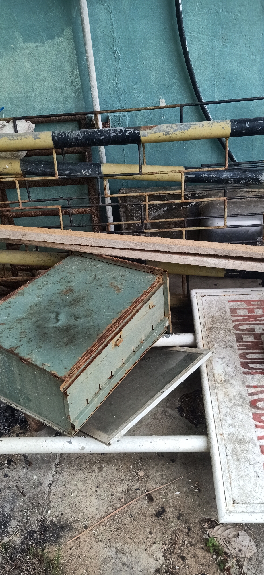 In Tambontunong, Indonesia, a collection of discarded metal objects rests against a turquoise-painted concrete wall. In the foreground, a faded green metal box with a rusty lid sits open, revealing a shallow interior. Beside it lies a white rectangular sign with red lettering, partially obscured. Behind these items, a jumble of rusty metal bars and grates are piled up. A white pipe runs horizontally across the bottom of the frame, and another white pipe, thicker and vertical, is visible against the wall. The ground is a mixture of concrete and dirt, littered with debris.