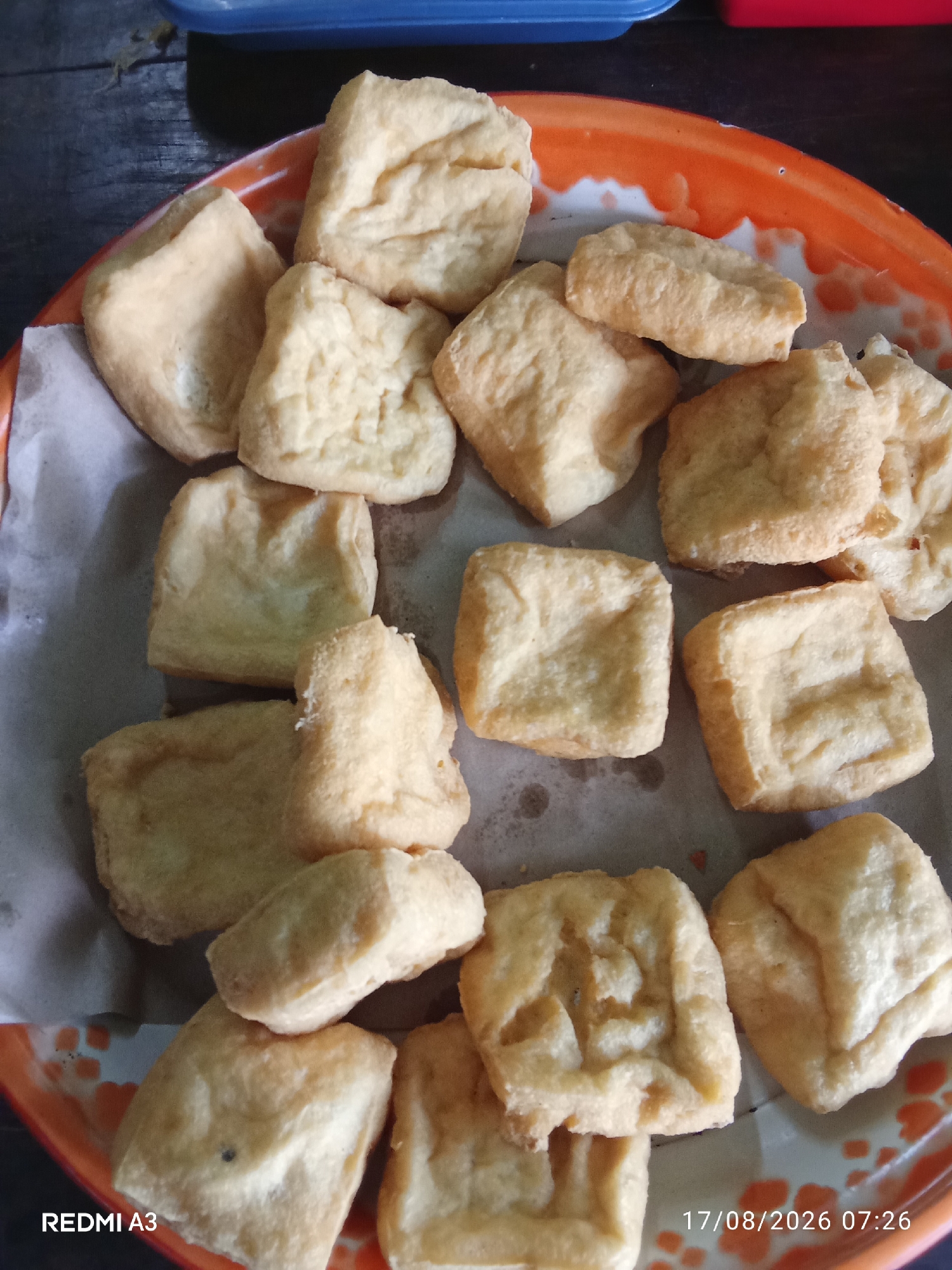 A close-up, top-down shot shows a pile of fried tofu pieces on a round, orange and white metal tray lined with white paper. The tofu is cut into irregular, bite-sized cubes, with a light golden-brown exterior and a slightly puffed interior texture. Some pieces are stacked atop others, forming a disordered heap in the center of the tray. The tray itself is slightly worn, with visible scratches and some orange paint chipped off along the rim, revealing a white base.
The background is dimly lit, with a dark wooden surface visible in the upper left corner, possibly a table. A portion of a blue container is also visible at the top edge of the frame, and a sliver of a red container is present to its right. The bottom of the image displays a timestamp "17/08/2026 07:26" and the text "REDMI A3" in white, indicating the device used for capture.
While the image is detailed, there are no people, animals, or any specific landmarks visible that would definitively place the scene within the city of Sanggrahan, Indonesia. The overall impression is a simple, informal presentation of food.