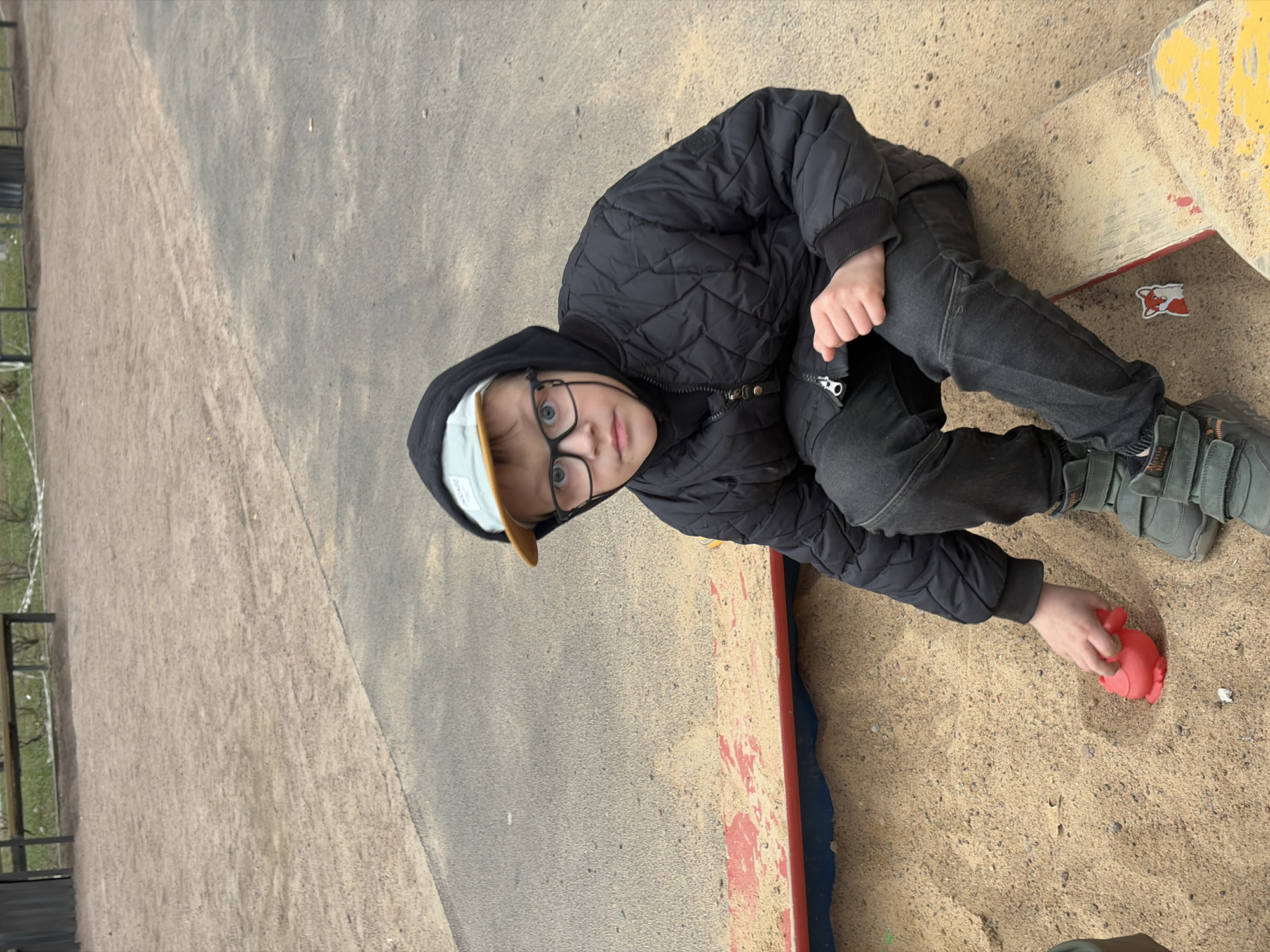 A young boy is playing in a sandbox in Saint Petersburg, Russia. He is wearing a black quilted jacket, black jeans, gray sneakers, and a light-colored baseball cap. He also has glasses on. The sandbox is bordered by a painted edge with red, yellow, and blue sections. A red toy is in the boy's hand within the sandbox. The sandbox is situated next to a gray surface, possibly concrete or asphalt, and a fence is visible in the background.