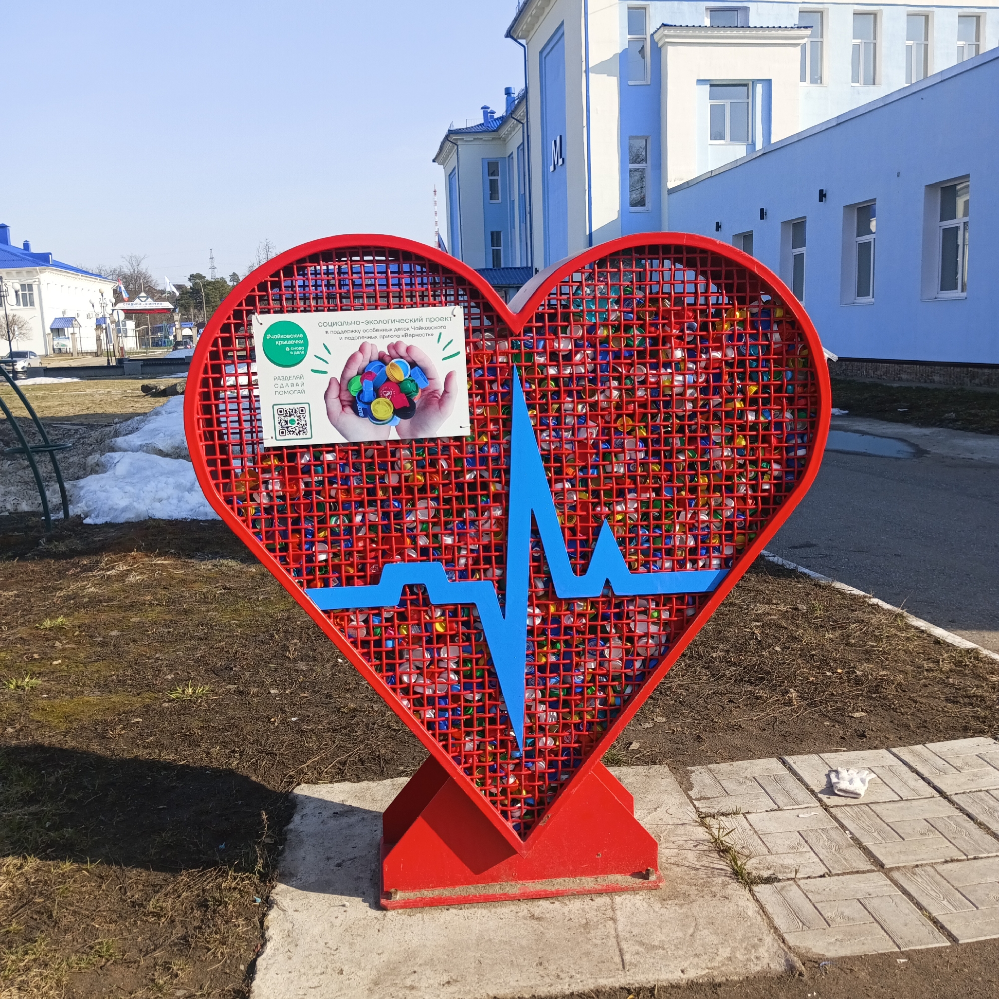 Here is a description of the image:
The image showcases a large, red heart-shaped structure filled with colorful bottle caps, standing on a paved area next to a patch of grass and a building. A blue, stylized heartbeat line runs horizontally across the center of the heart. Affixed to the structure is a sign with text and an image of hands holding bottle caps. Text on the sign identifies this as a social and ecological project in support of children and a local animal shelter in Chaikovsky, Russia. The sign includes the hashtags #ChaikovskyBottleCaps, and a call to action to "Separate, Hand Over, Help". The sign also includes a QR code. A white object resembling a cloth or paper is lying on the pavement near the structure. The building in the background has a light blue facade with white window frames. The overall scene appears to be outdoors on a sunny day, possibly in a park or public area within the city of Chaikovsky, Russia. Patches of snow are visible on the grass.