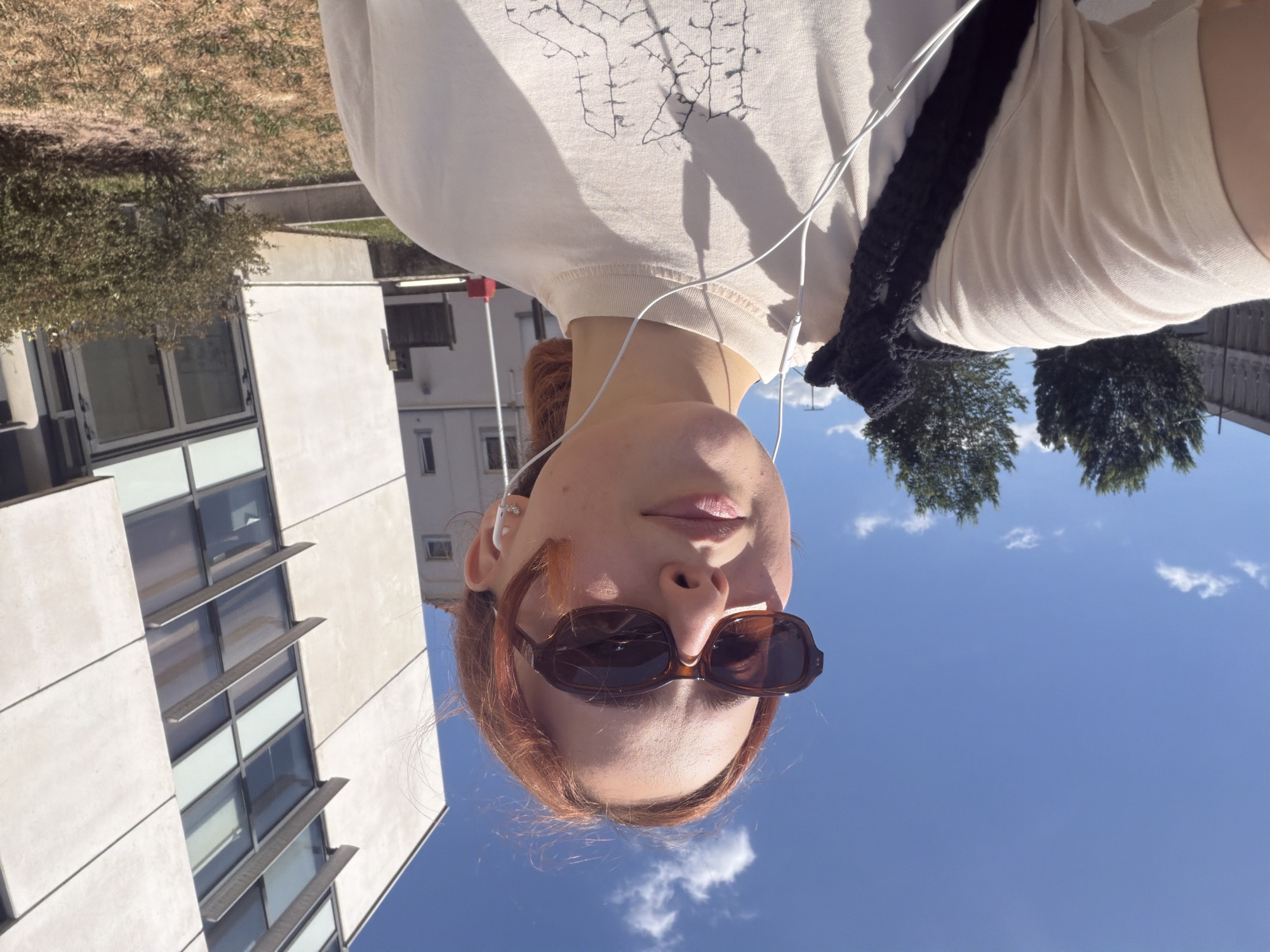 A young person with reddish hair is taking a selfie outdoors on a sunny day in front of a modern white building with glass windows. They are wearing sunglasses, a light-colored t-shirt with a graphic print, and white earbuds. A black knitted bag strap is visible over their shoulder. The background shows a clear blue sky with a few clouds, trees, and a concrete fence. The overall impression is one of a casual moment in an urban setting. It is not possible to definitively identify the city as Strasbourg, France from the visual information provided.