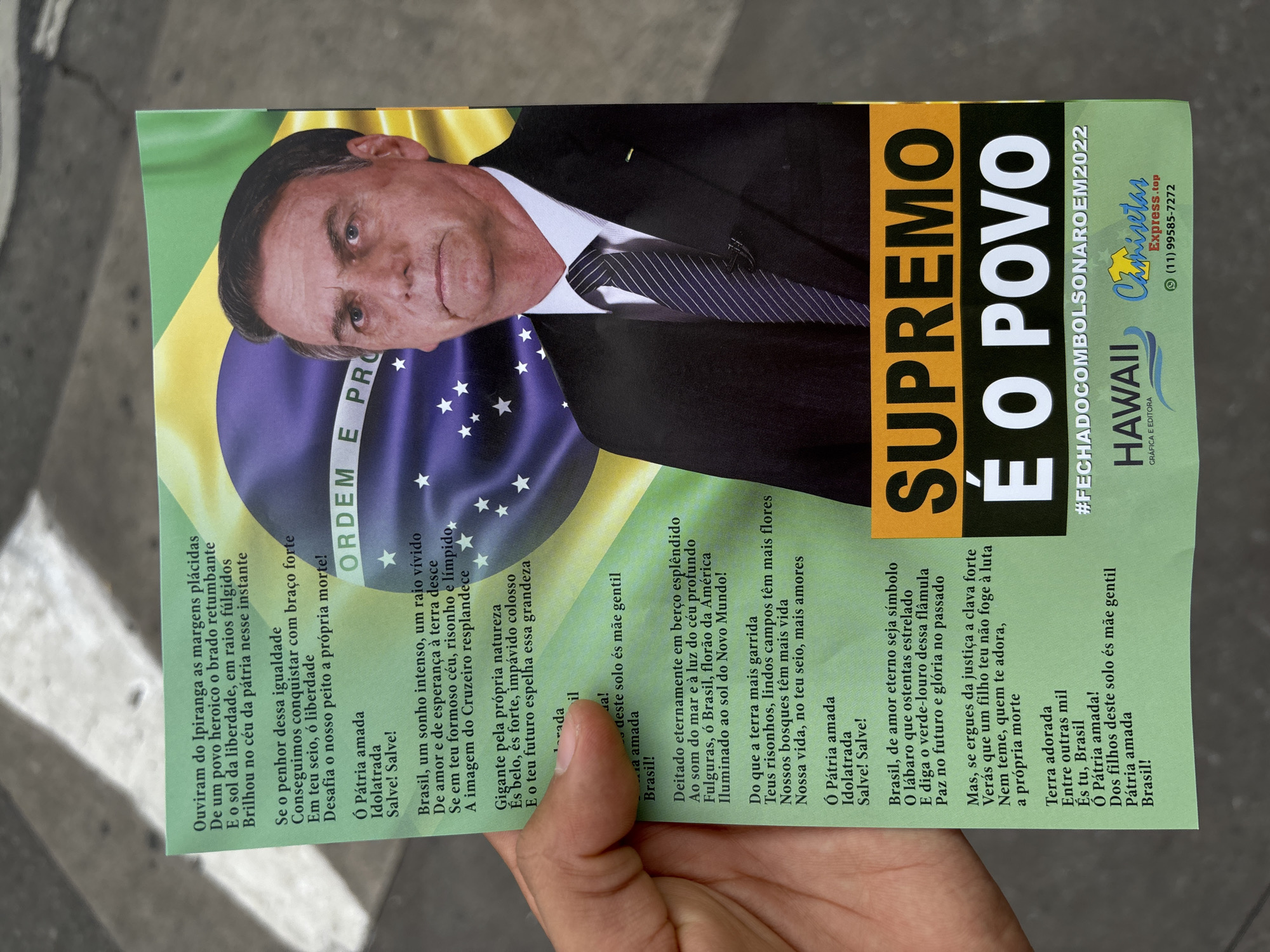 A printed political campaign flyer is held by a person's hand, against a blurred, light-colored background suggesting an outdoor or urban setting in São Paulo, Brazil. The dominant feature of the flyer is a portrait of Jair Bolsonaro, former president of Brazil, shown from the chest up, wearing a dark suit and striped tie, looking directly at the viewer with a serious expression. Behind his image is a depiction of the Brazilian flag, with parts of "ORDEM E PRO" (from "Ordem e Progresso") visible on the blue sphere.
Prominent text on the right side of the flyer reads "SUPREMO É O POVO" (The People are Supreme) in large yellow capital letters. Below this, a hashtag "#FECHADOCOMBOLSONAROEM2022" (roughly "Closed with Bolsonaro in 2022") indicates the campaign's political affiliation and timing. Further details include the names "HAWAII GRÁFICA E EDITORA" and "Chamisetas Express.top" with a São Paulo phone number "(11) 99585-7272". The left and bottom sections of the flyer are covered with verses from the Brazilian National Anthem, printed in smaller font. The flyer's design, content, and the São Paulo phone number firmly place its context within a Brazilian political campaign, likely related to the 2022 elections.