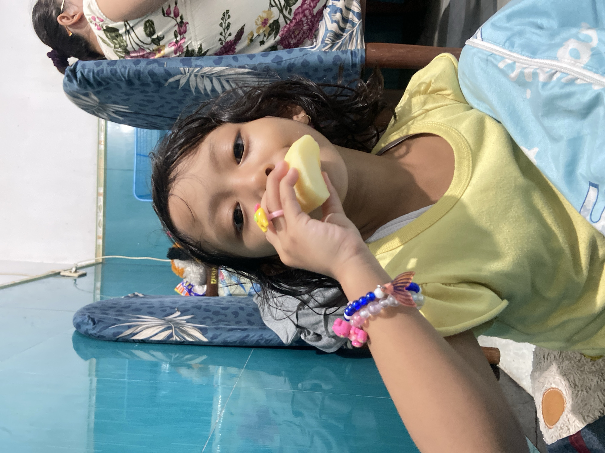 A young Asian girl is seen indoors, wearing a yellow shirt and holding a piece of peeled, light-yellow fruit, possibly an apple or pear, close to her mouth as if about to take a bite. She has a ring with a yellow charm on her finger and a colorful bracelet on her wrist. Her dark hair is slightly damp, and she is looking directly at the camera. Behind her, a woman is partially visible, wearing a floral patterned top, seated on a blue cushioned seat with a floral pattern. The background reveals a glossy blue-green tiled floor, a gray piece of cloth, and various objects including a small stuffed toy. This scene appears to be taken indoors, likely in a residential setting in Gelam, Indonesia.