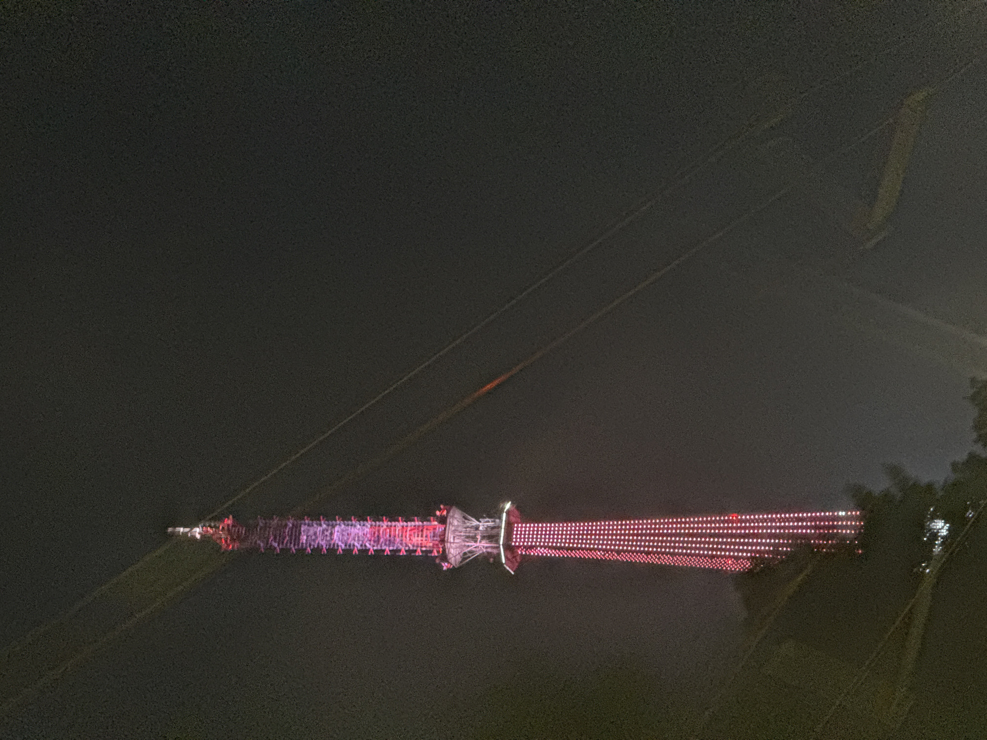 The image captures the illuminated Nizhny Novgorod TV Tower at night. The tower is brightly lit with pink and red lights, displaying a distinct pattern. Support cables stretch from the tower diagonally across the dark sky. Nearby, trees are partially visible. The sky is dark and appears to be somewhat foggy or hazy.