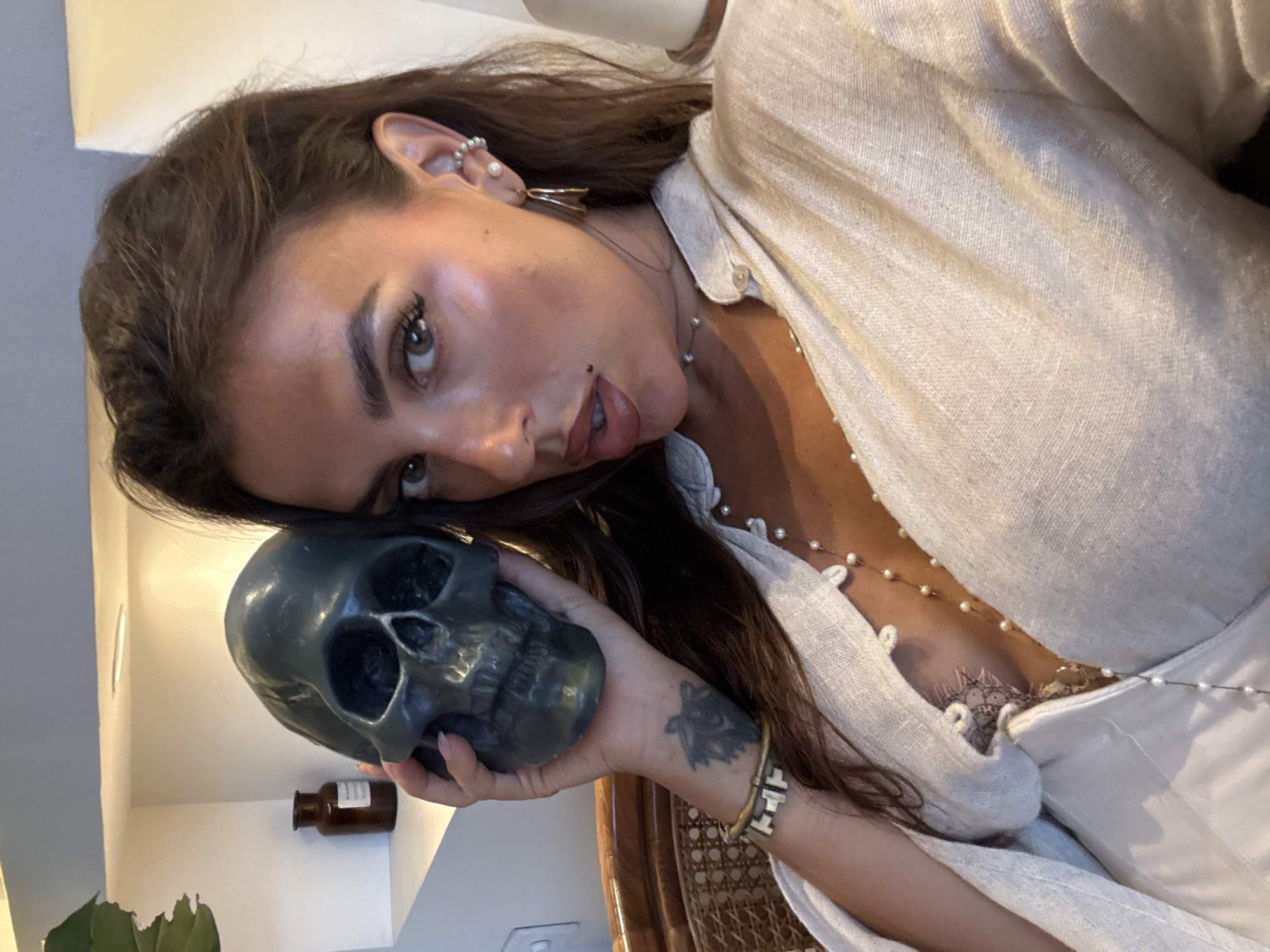 A young woman is posing indoors, holding a skull against her face. She has brown hair, light skin, and is wearing a light-colored linen shirt with a string of pearls. Her left arm is visible, showing a tattoo and a bracelet. The skull is dark gray. In the background, there is a small bottle on a shelf. The location is North Kuta, Indonesia.