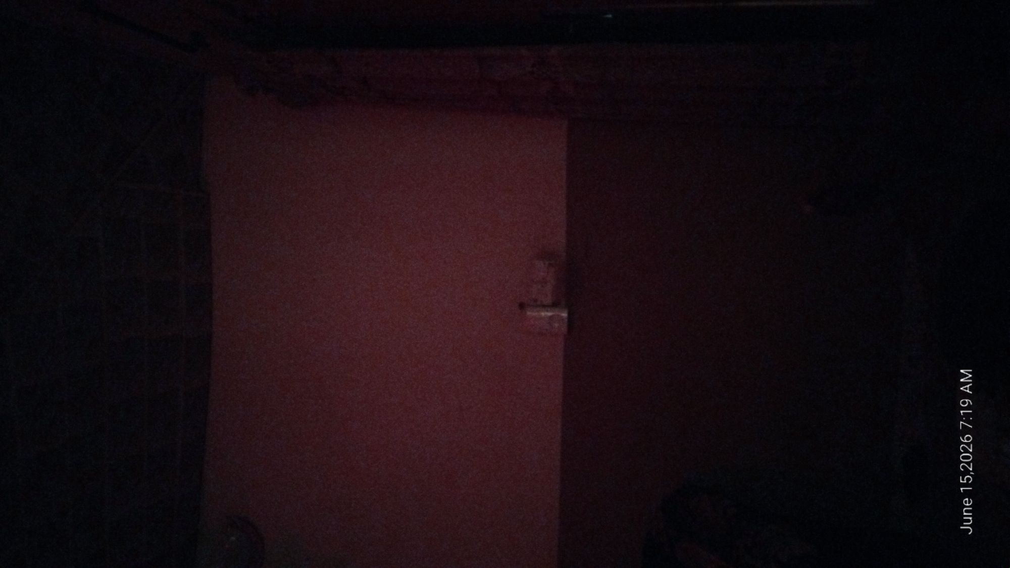 The image shows a dimly lit interior room with a dark ceiling and a pinkish-red wall. A dark brown lower section of the wall is visible at the bottom of the frame, with a dark object partially visible on the left. A small white electrical outlet or fixture is mounted on the pinkish-red wall. The lighting is very poor, obscuring many details. The date stamp in the lower left corner reads "June 15, 2026 7:19 AM". The location of Howayi, Nigeria, is not identifiable from the visual content of the image.