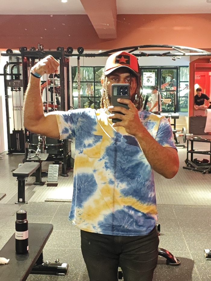 The image depicts a mirror selfie taken by an adult male in an indoor gym located in the city of Chongqing High-tech Zone, China. The man, with dreadlocks and a beard, is positioned centrally, holding a black smartphone with multiple camera lenses in his right hand to capture his reflection. He is wearing a blue and yellow tie-dye short-sleeved t-shirt, dark shorts or trousers, and a red baseball cap featuring a black patch with a red star. His left arm is flexed, showcasing his bicep, with a light blue wristband visible on his left wrist.
In the lower left foreground, a black flat workout bench holds a black insulated water bottle with a pink label. The gym floor is dark, textured rubber matting. The background reveals various gym equipment, including weight racks, benches, and machines, with "HAMMER STRENGTH" partially legible on one apparatus. Large windows or glass partitions in the background show glimpses of green foliage outside. Reflections in the mirror also show two other individuals in the gym: one person in a light-colored t-shirt in the mid-left background, and another person in a dark t-shirt sitting or crouching on the right. The upper walls and ceiling exhibit a reddish-orange hue, likely from artificial lighting, contrasting with lighter-colored walls elsewhere.
