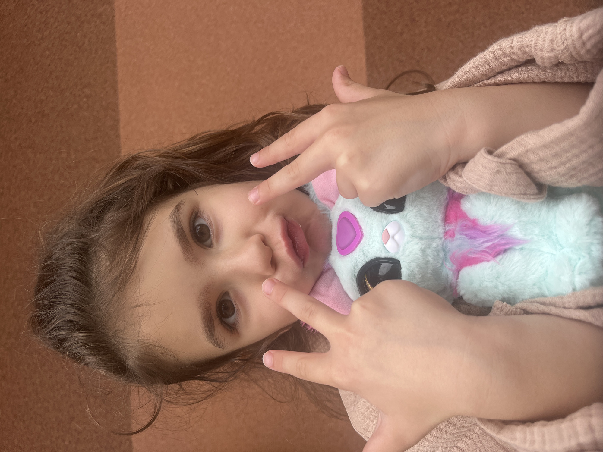 A young child, likely a girl of pre-adolescent age, is depicted in a close-up, chest-up portrait against a uniform reddish-brown background. She has long, dark brown wavy hair and dark brown eyes, which are directed at the camera. Her mouth is pursed into a playful pout. She wears a light pink or peach-colored knitted garment with a textured weave. Both of her hands are raised, each displaying a 'peace sign' or 'V' gesture with the index and middle fingers extended, positioned on either side of her face. Between her hands and torso, she holds a light blue or mint green plush toy featuring large black eyes, a white nose, pink-lined ears, a pink heart-shaped emblem on its chest, and a patch of multi-colored (pink, purple, blue) fur. There are no discernible details within the image to indicate the specific geographical location of Onokivtsi, Ukraine.