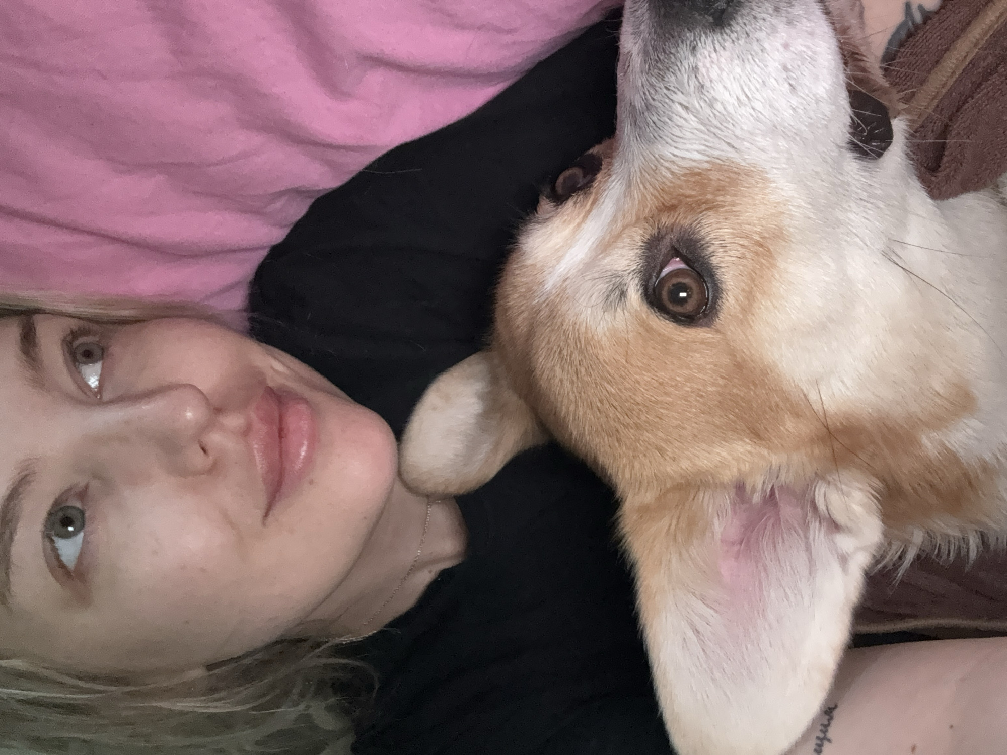 A selfie-style close-up photograph shows a fair-skinned woman and a dog lying down. The woman, positioned on the left side of the frame, has blonde hair, light-colored eyes, and her face is slightly angled upwards. Her lips appear glossy. To her right, filling the majority of the frame, is the head of a tan and white dog. The dog's head is oriented upside down relative to the woman's. The dog's brown eyes are visible. The scene appears to be indoors, with a pink fabric visible in the upper left corner, possibly a blanket or pillow. The woman is wearing a black top, and a thin necklace is visible around her neck. The dog is wearing a dark-colored collar. Part of a tattoo is visible on the woman's arm. No specific information suggests the location is Borzya, Russia.
