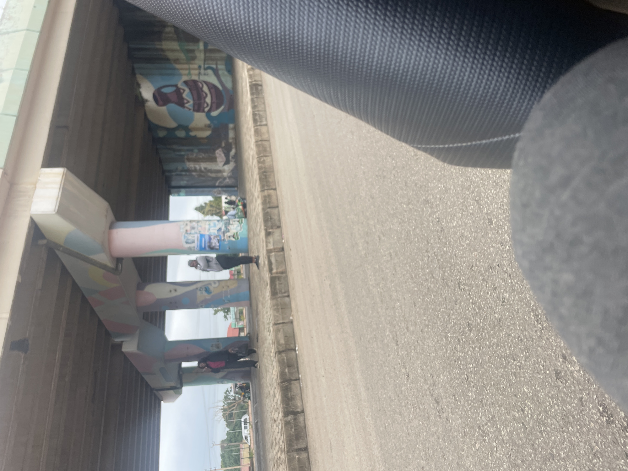 The image depicts an underpass in Jos, Nigeria, with several individuals visible along the pedestrian walkway. The underpass pillars are adorned with colorful abstract paintings and murals, including a depiction of a calabash. Two women are standing on the walkway, one in a pink jacket and dark pants, carrying a bag, and another in a grey hoodie and dark pants. A few other figures are also present further down the walkway. The scene appears to be a transit point, possibly a bus stop, given the presence of people waiting. The road surface in the foreground suggests a busy urban environment. A portion of a vehicle's seat or interior is visible in the lower right corner of the frame, implying the photograph was taken from a moving vehicle.