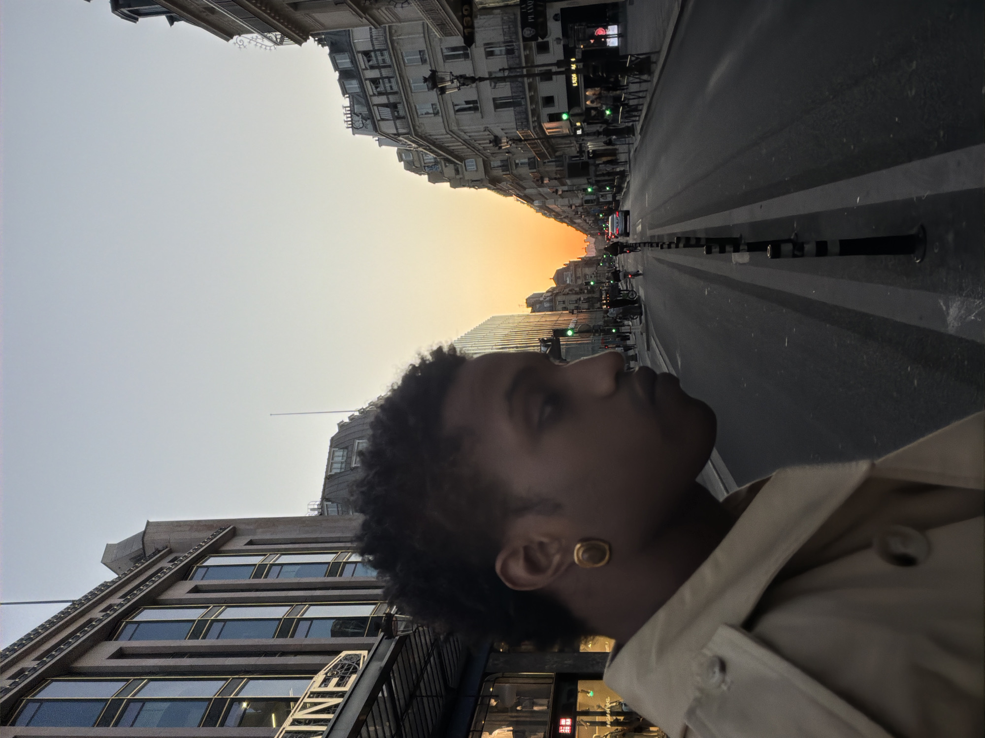 The image presents an urban street scene in Paris, France, captured during either sunrise or sunset, characterized by a vivid orange and yellow glow on the horizon that fades into a lighter, cooler sky above.
In the foreground, a person with dark, curly hair is depicted in profile, looking contemplatively towards the distant horizon. They are wearing a light-colored, possibly beige, jacket or coat and a prominent gold-colored stud earring.
The street extends into the distance, flanked on both sides by tall, multi-story buildings with classic European architecture, including detailed facades and balconies. On the right side of the street, street lights, traffic lights (some showing green), and bollards are visible. Distant pedestrians and vehicles suggest a lively city environment.
Notable visible text includes "MONACO" and a partially inverted "CHANEL" on building signs. A vertical inscription "NIVE" (likely part of a brand name) is seen on a building near the foreground subject. Further down the street, a "UNIQLO" sign with additional red characters, possibly in an Asian script, is visible on a shopfront. The scene conveys a tranquil moment amidst the bustling city.