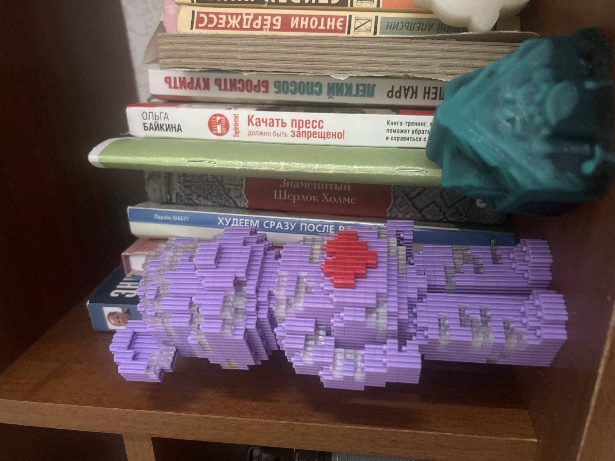 The image shows a close-up view of a shelf in a room, likely a home library or study. The shelf is made of wood and has a dark brown finish. On the shelf are several books, a purple brick-built bear figurine with a red heart on its chest, and a teal-colored object that appears to be a wax melt or soap. The bear figurine is standing upright and is made of small, interlocking bricks. It is detailed with a red heart shape in the center of its chest. The teal object is irregularly shaped and textured, resembling a rock or a piece of abstract art. The books are of various sizes and colors, with some titles visible in Russian. The overall scene suggests a personal collection of items displayed on a shelf. The location of Pskov, Russia is not indicated by any visual cues in the image.