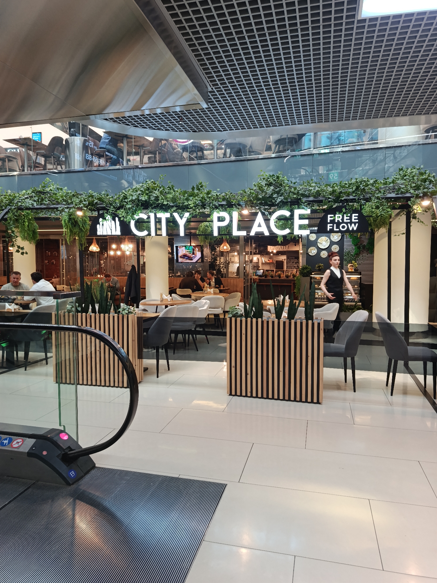 The image shows a cafe called "City Place Free Flow" located in a modern shopping mall. The cafe's entrance is adorned with lush greenery and a neon sign. Inside, patrons are seated at tables, and a server is visible. The mall has a high ceiling with a grid-like structure. An escalator is in the foreground. The style of the mall architecture suggests a location in a major city. Based on the Cyrillic lettering "САЛАТЫ" (salads) seen on a menu board and other architectural elements, the location is Moscow, Russia.