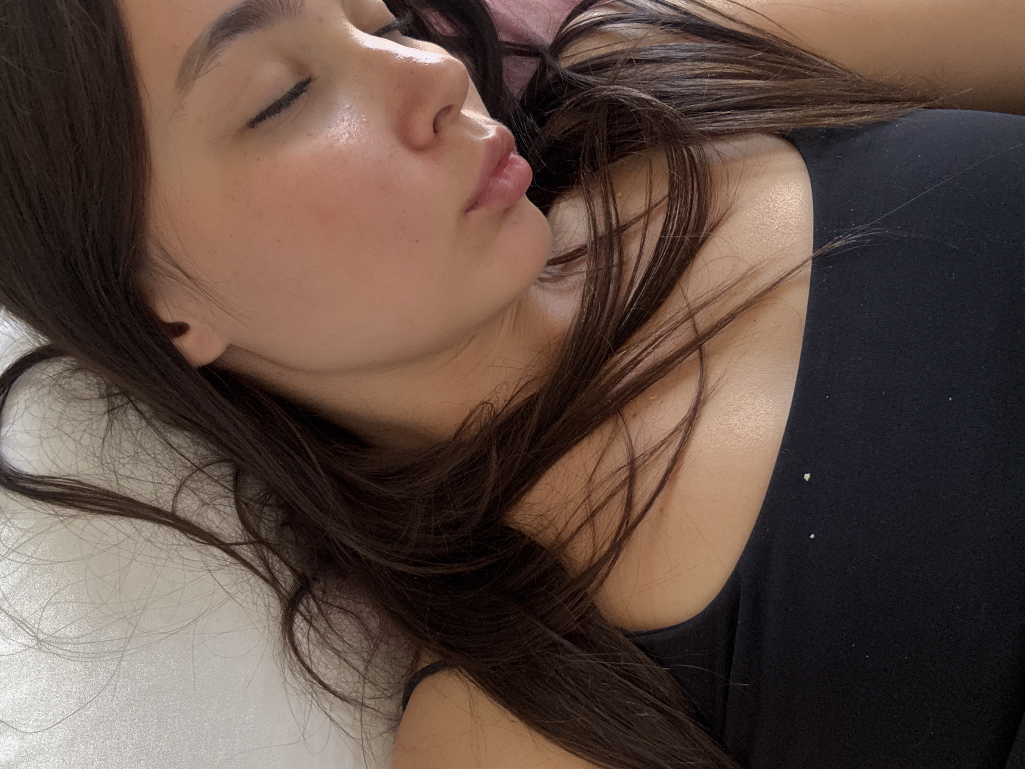 A woman with dark brown hair lies on her back on a white pillow. Her eyes are closed. She wears a black top. The location is Unknown, Unknown.