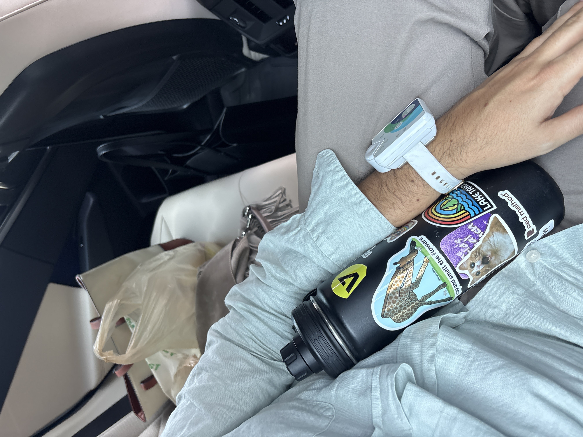 The image shows a person sitting in a car. The person is wearing a light blue button-down shirt and a white device is attached to their wrist. A black water bottle with several stickers is resting on their arm. The stickers include images of a giraffe, a cat, a logo with a yellow "V," and text reading "LAKE TAHOE" and "Red method." Shopping bags and a purse are visible on the seat next to the person. The interior of the car is primarily black and white. The location is not specified, therefore, the location in the city of Mexico City, Mexico is unknown.