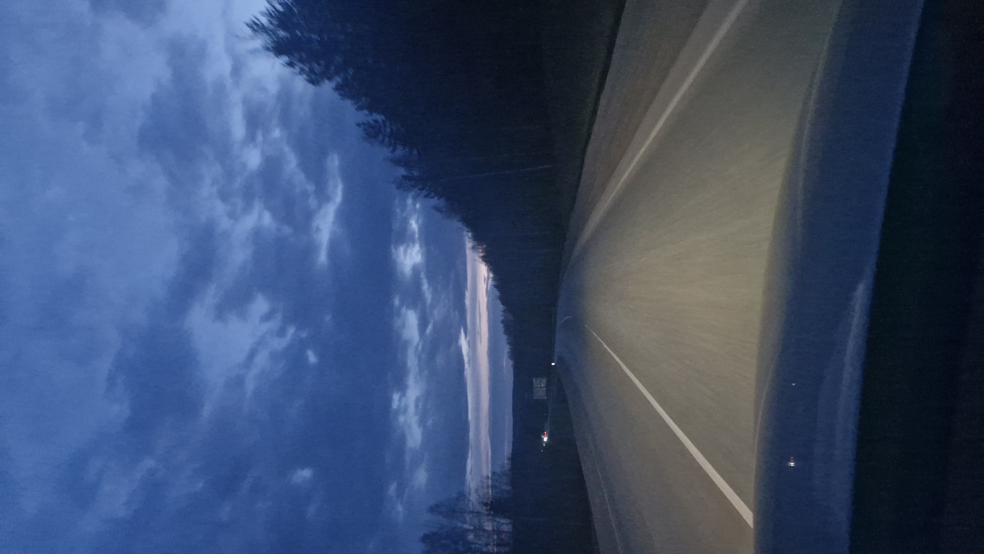 The image, oriented horizontally, depicts a road scene at twilight or dusk in Selishcha, Russia. The perspective is from inside a moving vehicle, looking forward along a paved road.
The sky, occupying the upper left portion of the frame, is a deep, muted blue-grey, filled with indistinct, textured clouds, suggesting an overcast or partly cloudy evening. Along the horizon, a slender, horizontal band of soft, light pinkish-purple hues indicates the last light of the setting sun or the earliest light before sunrise.
The road itself, made of asphalt, extends from the lower right foreground into the distance, appearing to curve gently to the right. A distinct white line marks the right edge of the visible road. The road surface in the foreground and mid-ground is illuminated by a soft, warm light, likely from the vehicle's headlights, which casts a gentle glow.
To the left of the road, a dark, dense silhouette of trees lines the horizon, indicating a heavily forested area. In the far distance, a small, subtle red light is visible, possibly belonging to another vehicle or a distant object. No people are visible, and there is no discernible text. The scene conveys a quiet, contemplative atmosphere of travel through a natural landscape during the transition from day to night.