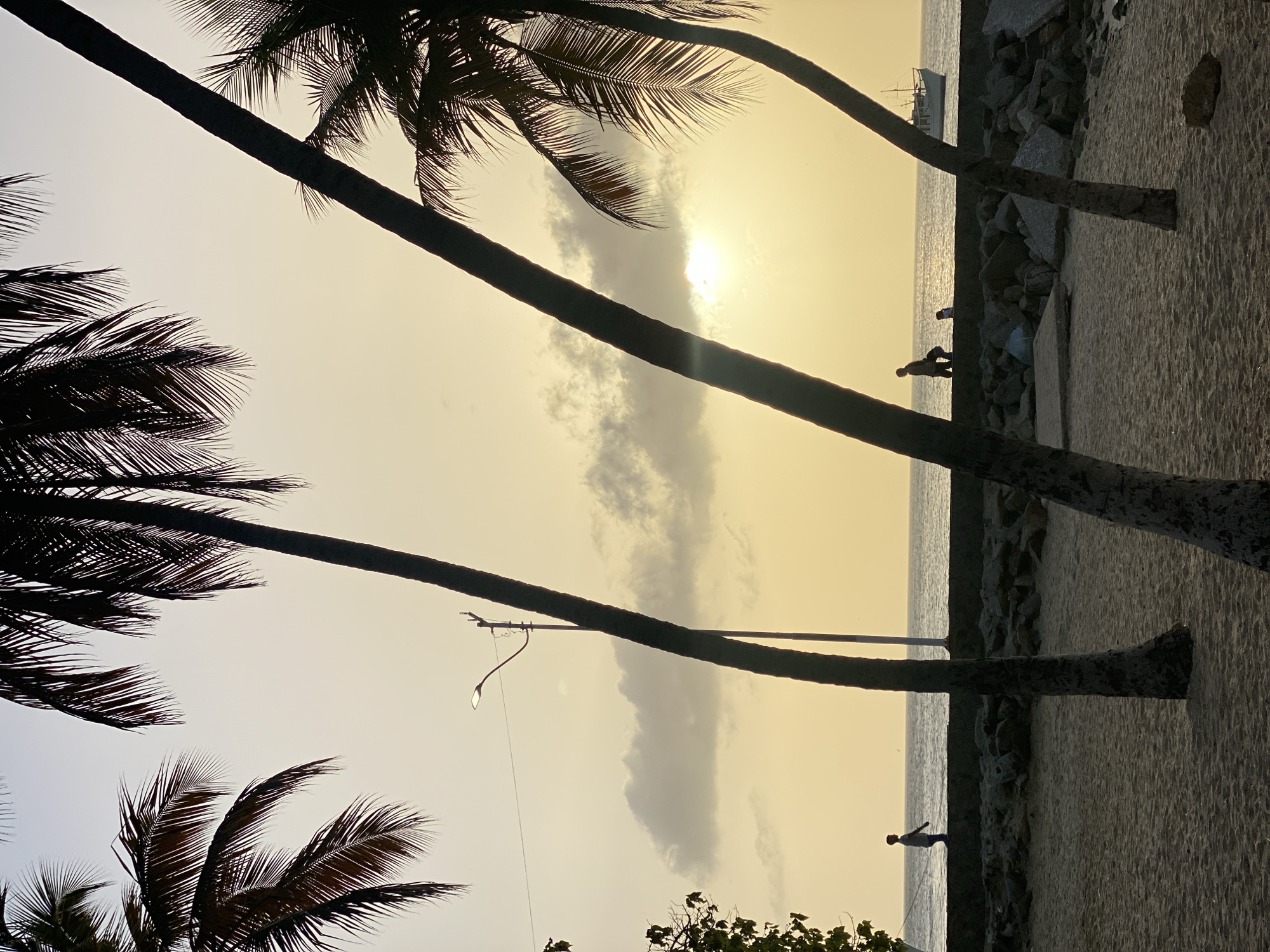 The image depicts a beach scene at sunset in Juan Griego, Venezuela. Silhouetted palm trees frame the view of the sky and ocean. The sun is visible through a break in the clouds. A breakwater separates the sand from the water. A person is standing on the breakwater. Another person walks along the beach. A boat is visible in the distance. A street light is visible among the palm trees.