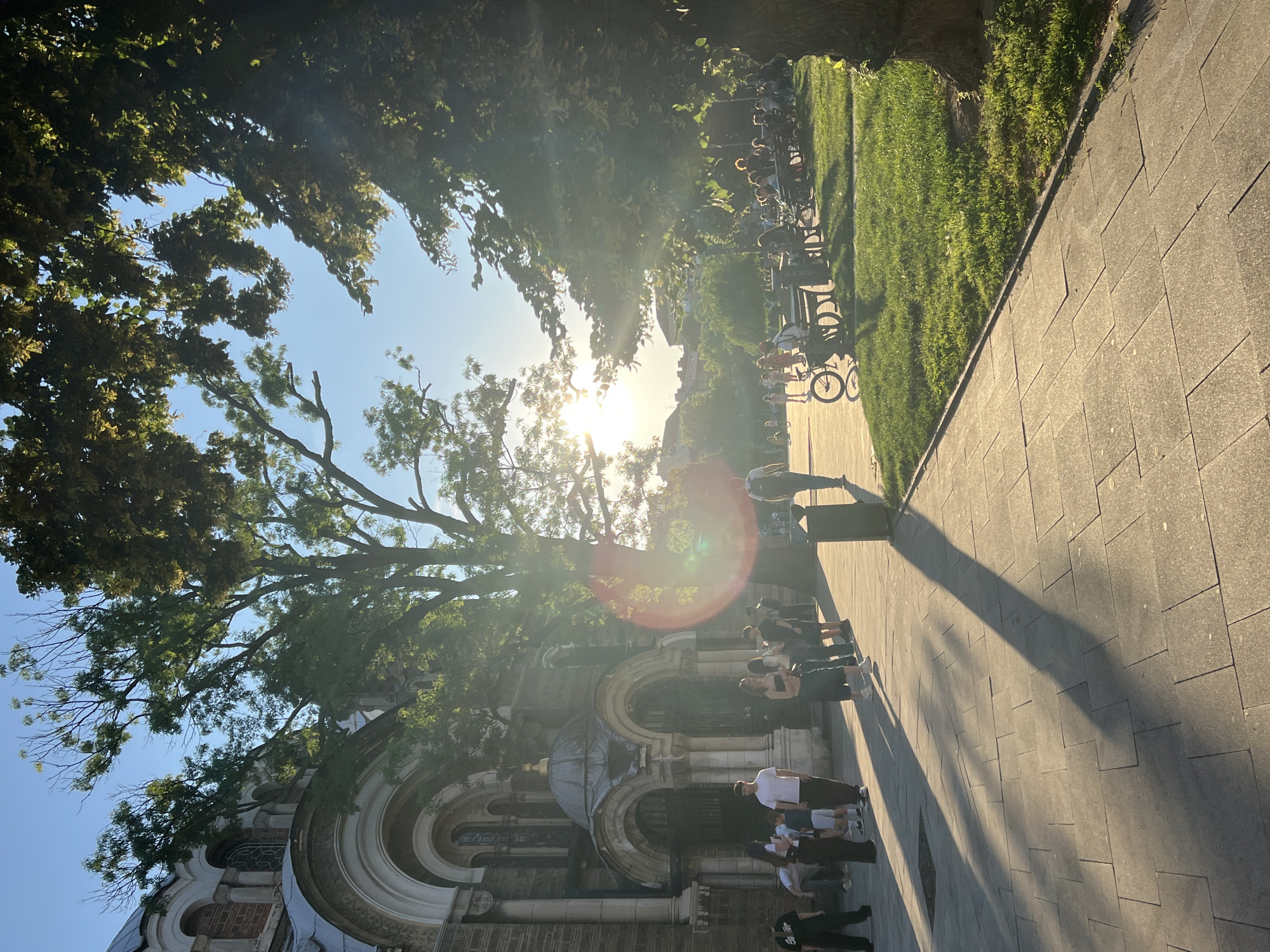 The image depicts a sunny scene outside a church, likely in Sofia, Bulgaria. The architecture suggests a religious building with arched entrances and a dome. Several people are visible, some standing in a line near the church entrance, while others are seated on benches in a grassy park area in the background. One person is on a bicycle. The sun is low in the sky, casting long shadows across the paved walkway. Trees with green foliage are prominent, framing the scene and dappled sunlight filters through their leaves. The atmosphere appears relaxed and communal, with people enjoying the warm weather.