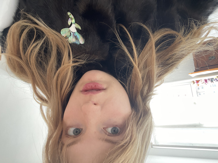 The image depicts a young Caucasian female in a close-up, chest-up shot, set within an indoor environment in the city of London, United Kingdom. She possesses long, wavy blonde hair with bangs, blue eyes, and slightly parted lips, looking directly at the viewer with an attentive expression. She is wearing a dark, possibly fur or faux fur, garment, adorned with an iridescent, abalone-shell-like floral brooch pinned to her left chest. The background reveals a brightly illuminated room with a large window to the left, through which a blurred view of an external urban or residential building is visible. Several small, colorful figurines are arranged on the windowsill below the window. A section of a wooden dresser or cabinet is partially visible to the far left of the frame.