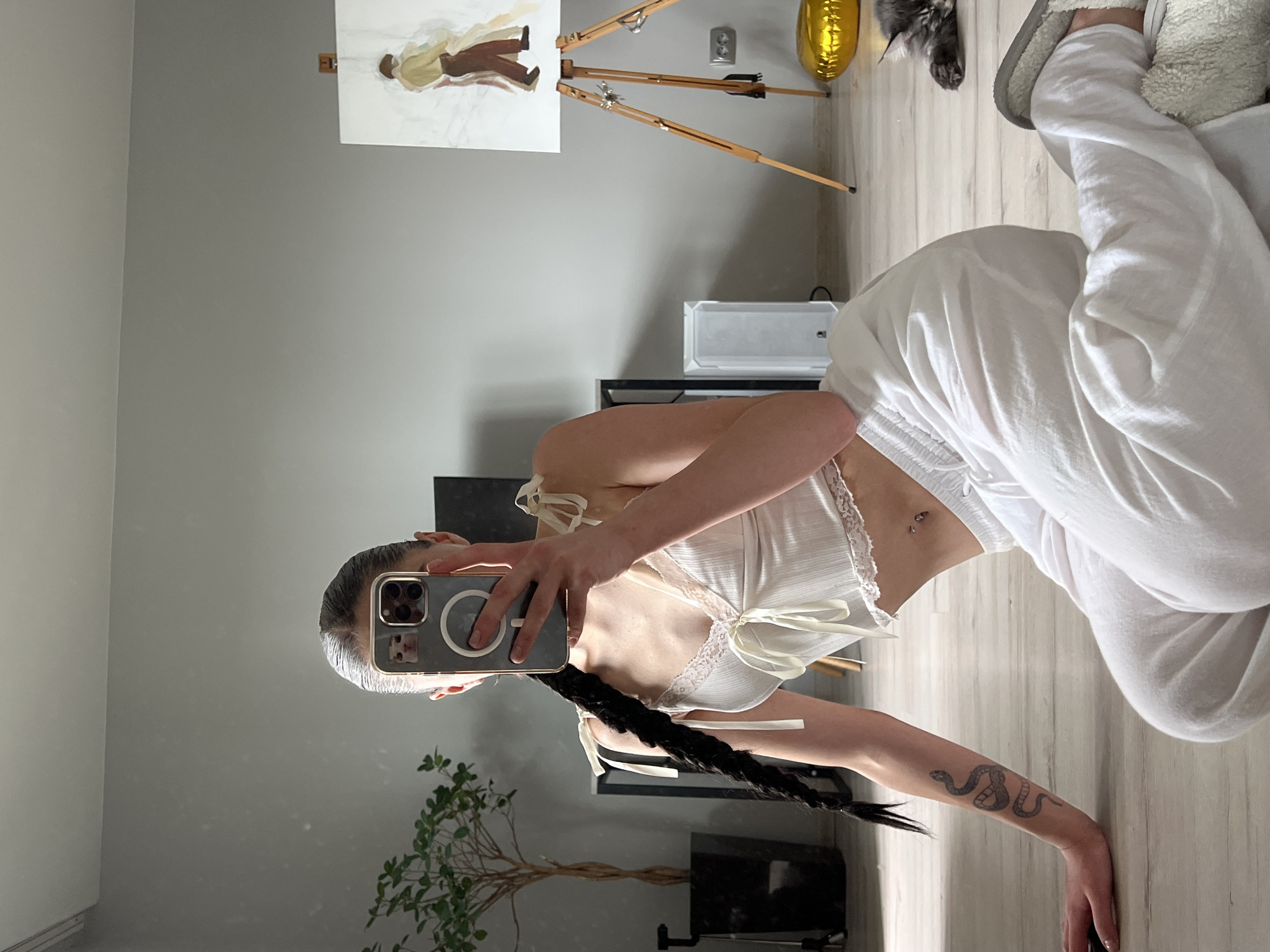 The image shows a young woman with dark hair styled in a long braid, taking a selfie in a room. She is wearing a white lace-trimmed tank top and white linen pants. A snake tattoo is visible on her left arm. She is sitting on a wooden floor, with her legs crossed. In the background, there is a plant in a black pot, a white speaker, a painting on an easel, a golden balloon, and a grey cat sleeping on the floor. The room appears to be a living space. The overall impression is casual and domestic.
