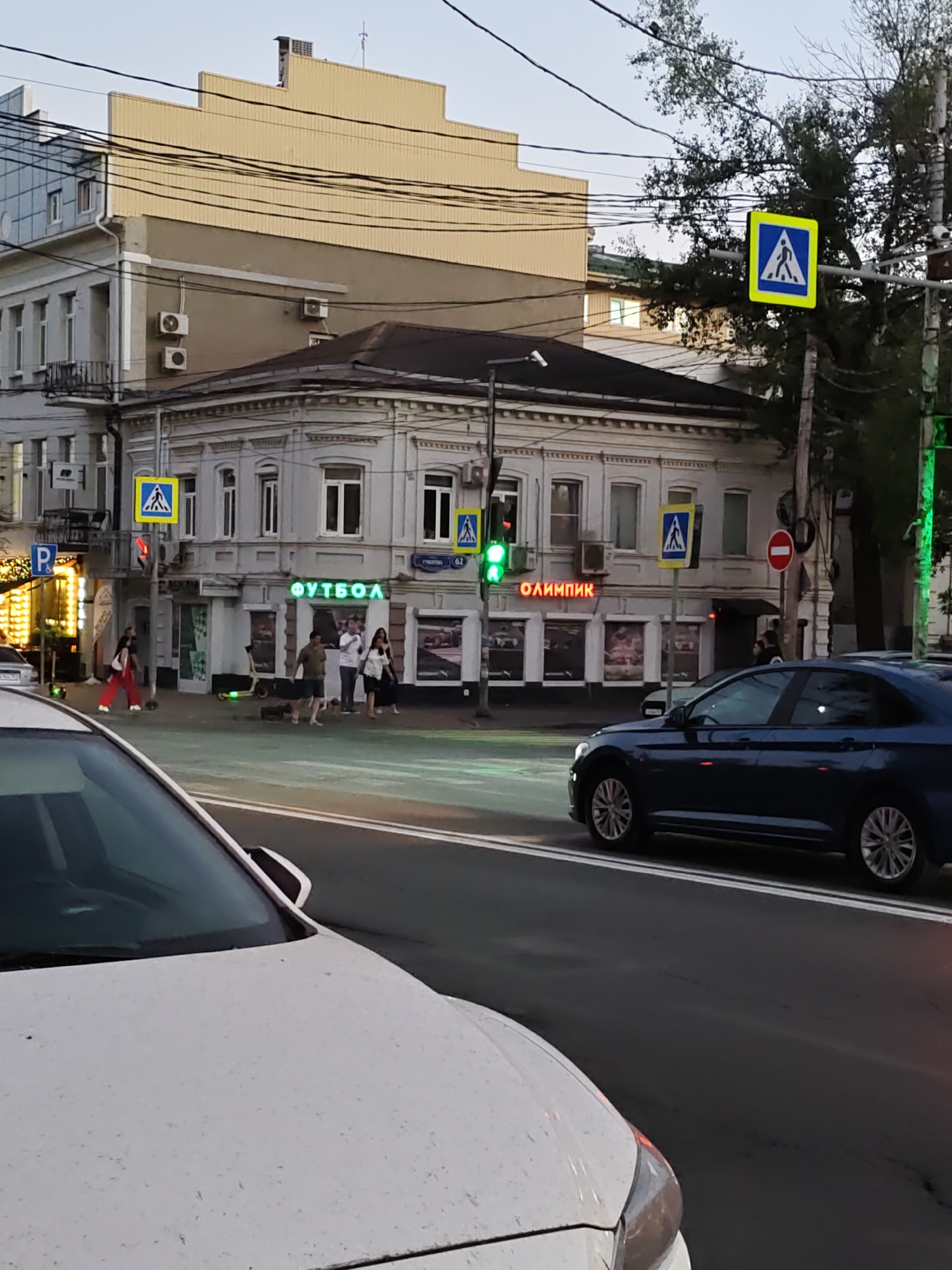 The image depicts a street scene in Rostov-on-Don, Russia. In the foreground, the hood and windshield of a white car are visible. The street is paved with asphalt and marked with white lines. A dark blue sedan is driving on the road to the right of the white car.
In the background, a white multi-story building with ornate architectural details stands. Two neon signs are mounted on its facade; one reads "ФУТБОЛ" (Football) in green, and the other reads "ОЛИМПИК" (Olympic) in red. Several people are walking on the sidewalk in front of the building. A pedestrian crossing sign with a blue background and a white figure of a person walking is visible on a pole. Traffic lights are also present.
Further back, a beige multi-story building with a stepped roofline is visible, with electrical wires running across the scene. Trees with green foliage are also present, and the sky is a pale blue, suggesting it might be late afternoon or early evening. The overall scene is typical of an urban environment with commercial establishments and pedestrian activity.