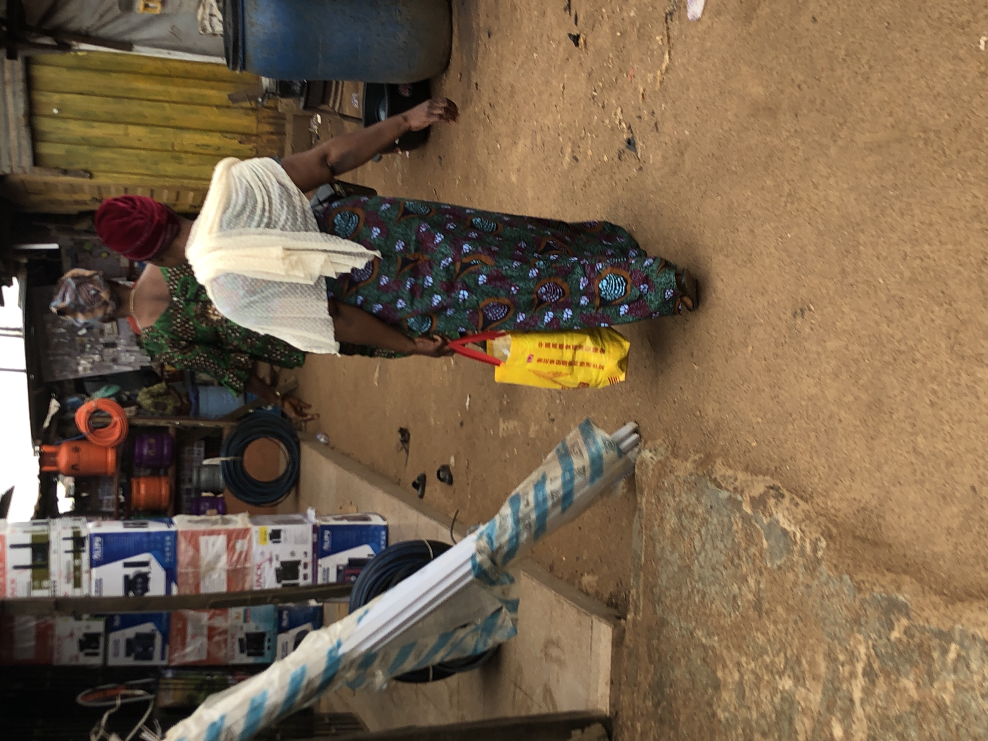 The image depicts a street scene in Ijoko, Nigeria. In the foreground, a woman wearing a red headscarf, a white sheer scarf draped over her shoulders, and a long, colorful patterned dress walks away from the camera. She is carrying a bright yellow shopping bag in her left hand. To her left, a long, white object wrapped in blue and white plastic sheeting leans against a stall. Behind the woman in the foreground, another woman wearing a green and black patterned dress and a colorful headwrap walks away. The environment includes stalls with stacked boxes, a large blue barrel, and orange gas cylinders. The ground is dirt.