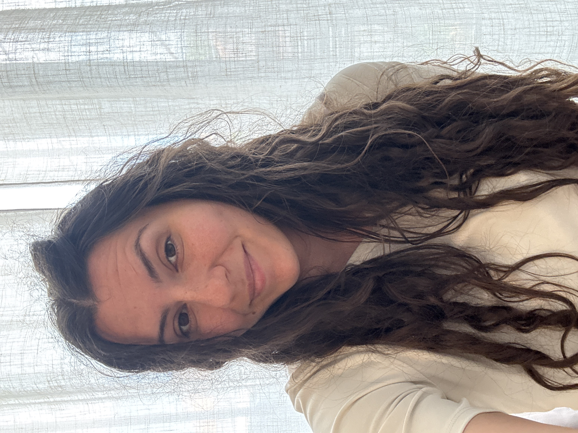 A woman with long, wavy brown hair is smiling towards the camera. She has light skin with freckles. She is wearing a light-colored shirt. The background features a sheer white curtain. Location: Unknown, Unknown.