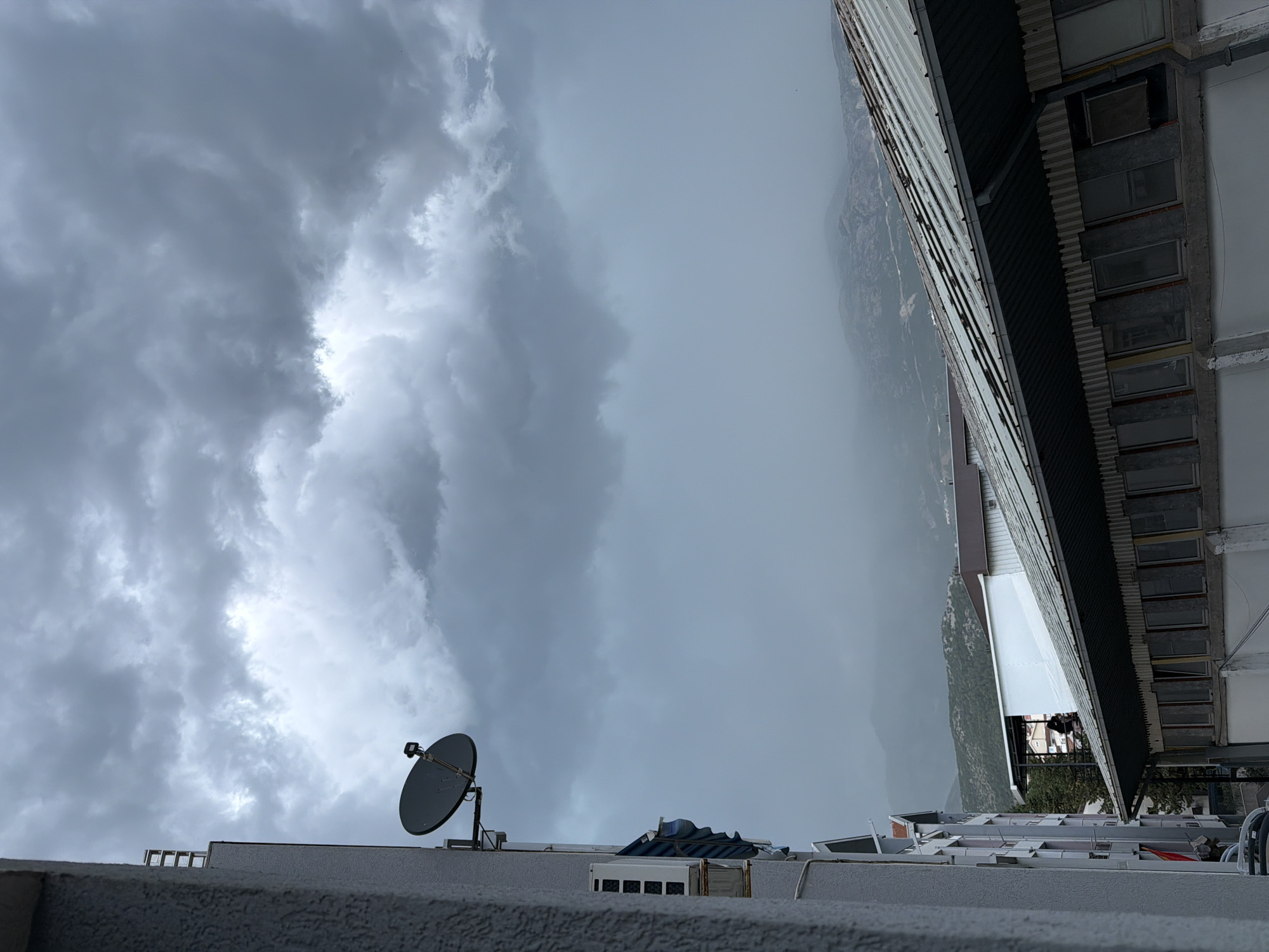 The image displays a partially rotated view of a cityscape and cloudy sky. The foreground shows the edge of a building with a textured wall, and on top of it, a satellite dish is visible. Further back, another building with a metallic roof and multiple windows is present, overlooking a hazy landscape of mountains. The sky is filled with dramatic, dark gray clouds, suggesting impending or ongoing rain. The lighting indicates it is daytime, but the overcast conditions create a somber atmosphere. There is no discernible text within the image.