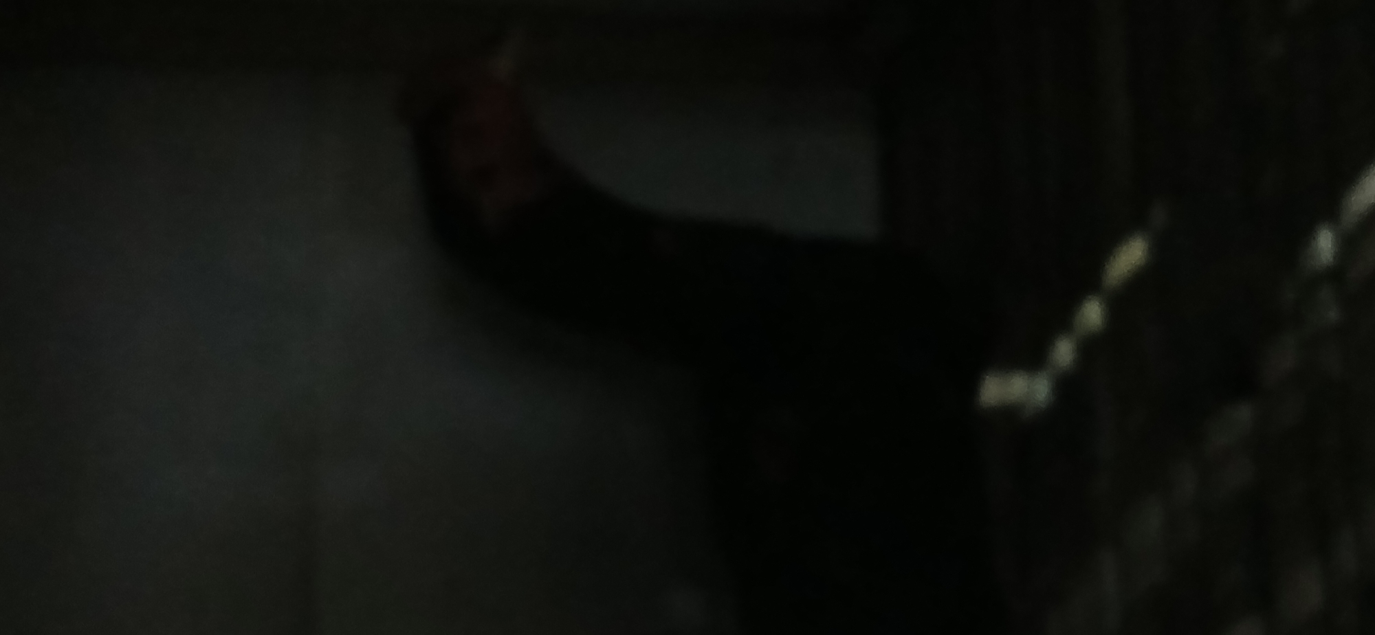The image depicts a dimly lit, indoor scene. A person is partially visible in the upper right quadrant, hunched over and appearing to be smoking. The environment is dark and nondescript, with no distinguishing features to suggest a specific location within Demangan, Indonesia, or elsewhere. The foreground shows a textured surface, possibly a carpet or rug, with some faint, light-colored linear elements. The overall impression is one of low light conditions and a lack of clarity.