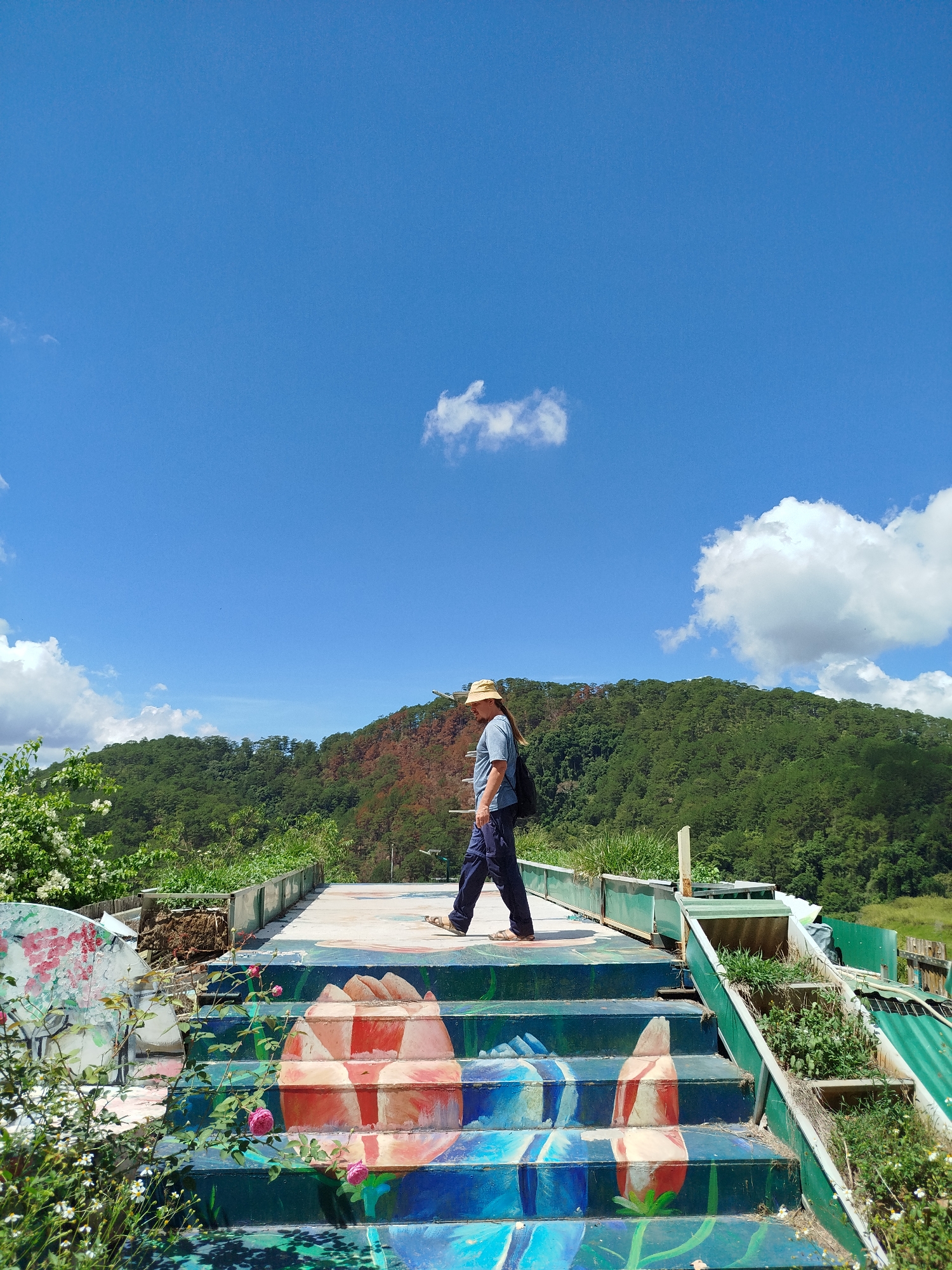A person wearing a light blue shirt, dark pants, sandals, and a straw hat walks across a platform with painted steps in an outdoor setting, possibly Da Lat, Vietnam. The steps are decorated with an abstract floral design, predominantly in shades of blue, green, white, and red. The platform appears to be a raised structure, and to the right, there are trough-like containers filled with plants. The surrounding environment includes lush greenery, possibly a hillside, and a bright blue sky with scattered white clouds. The scene suggests a tourist location or a unique architectural element in a natural setting.