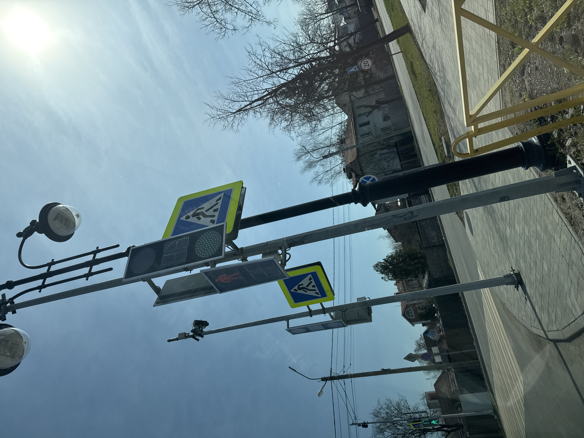The image shows a street scene in Novo-Nikol'skoe, Russia. The view is angled upwards, focusing on traffic infrastructure. There are pedestrian crossing signs with yellow borders and blue backgrounds, along with traffic lights displaying red. There are street lamps and a surveillance camera. The background includes buildings, trees with bare branches, and a sidewalk with a yellow railing. A "20" speed limit sign is also visible. The sky is light blue with bright sunlight.