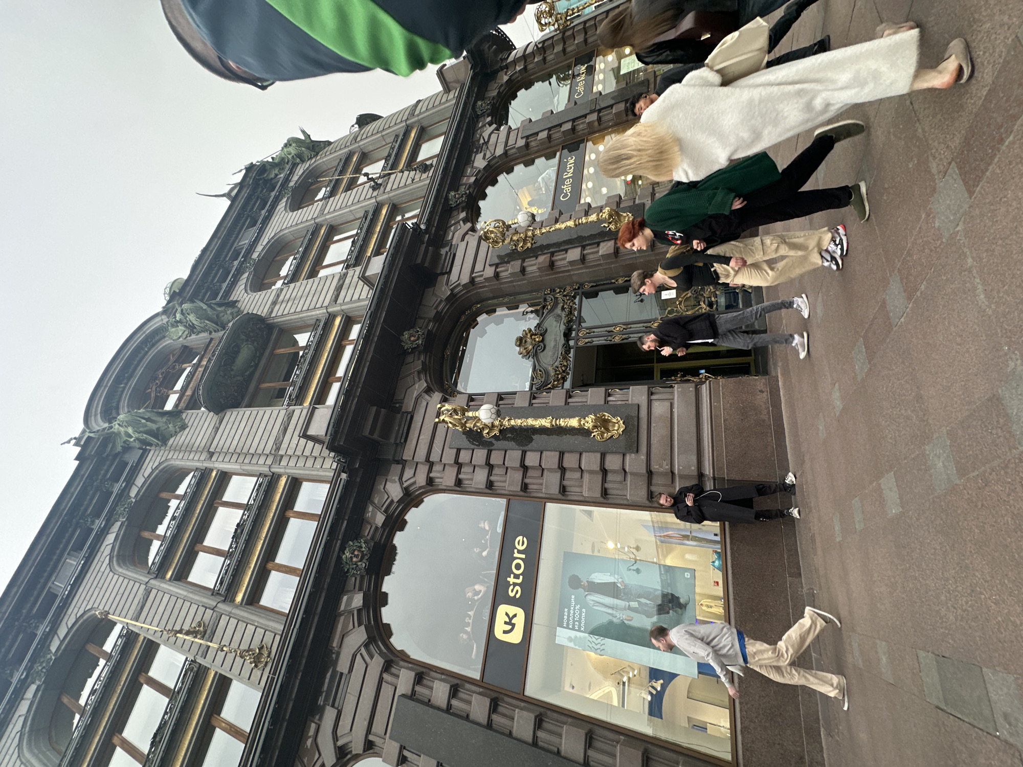 The image shows a street scene in Saint Petersburg, Russia, featuring the historic Eliseyev Emporium building. The Emporium is a large, ornate building with multiple floors, decorated with architectural details such as arched windows, bronze embellishments, and sculptures. A "VK Store" is visible on the ground level. People are walking along the sidewalk in front of the building. One man is walking in a beige set, and another is dressed in black and leaning against the building. Further down the sidewalk, there are more pedestrians, including a person wearing a white coat and another in a green sweater. The sky is overcast.