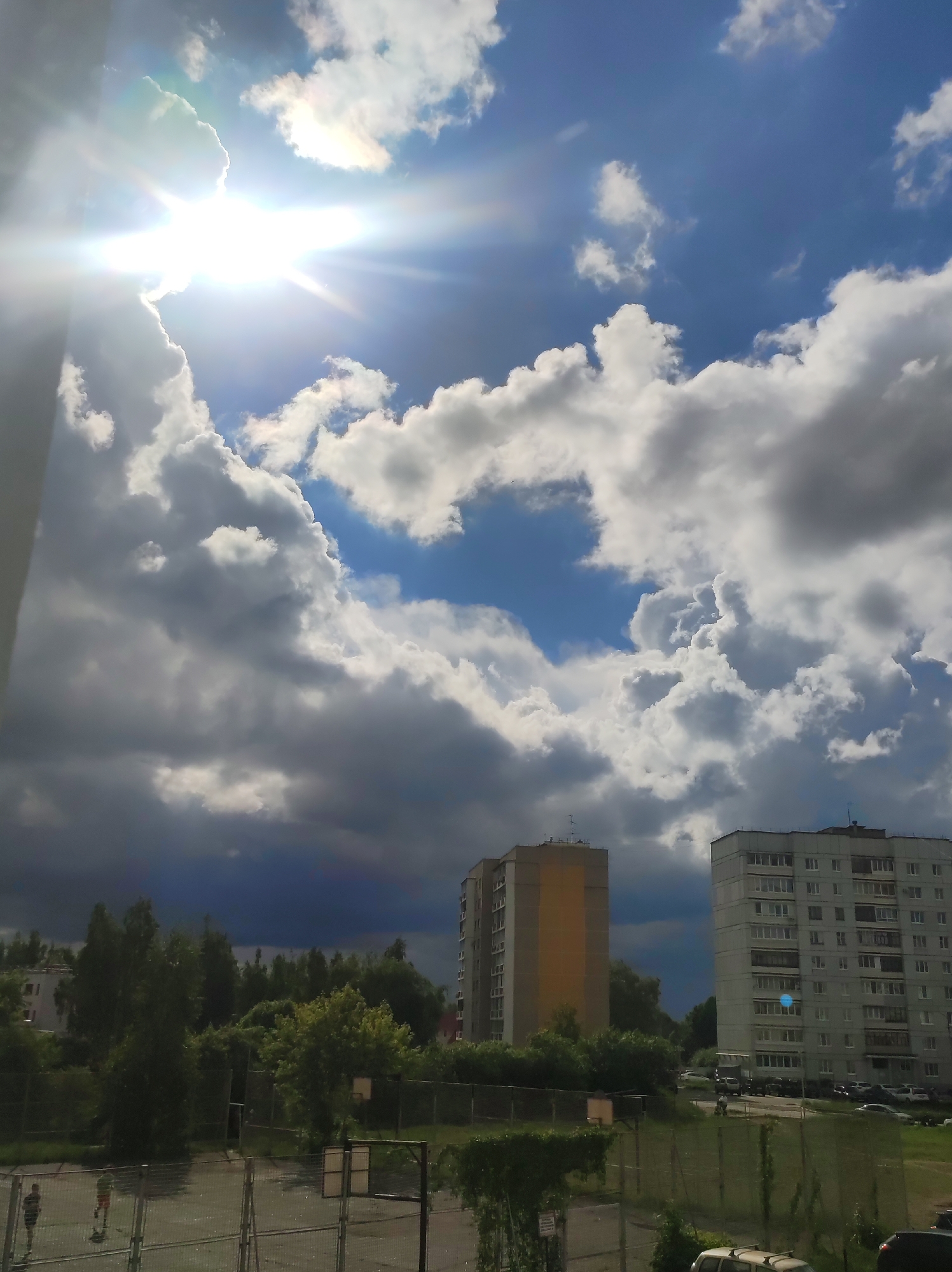 This image presents a vibrant and dramatic outdoor scene, likely captured from an elevated perspective overlooking a residential area in Chaikovsky, Russia. The sky is a dominant feature, showcasing a dynamic interplay of bright sunshine, clear blue patches, and expansive cumulus clouds ranging from brilliant white to deep, stormy grey. A powerful sunburst emanates from the upper left, casting intense light and lens flare across the scene.
In the middle ground, two multi-story apartment buildings stand prominently. The structure on the left is lighter-toned, possibly beige or yellow, while the building on the right is grey-white, both featuring numerous windows and balconies. Lush green trees and vegetation fill the spaces between the buildings and extend into the foreground.
The foreground reveals an outdoor sports court, possibly a basketball court, enclosed by a wire mesh fence. Two individuals are visible on the court, engaged in an activity that appears to be playing or walking; one wears a green/red shirt, the other darker clothing. The court surface shows reflections, suggesting it might be wet from recent rain. Several parked cars can be seen along a road behind the right apartment building.
The scene depicts daytime, likely late morning or afternoon, characterized by a striking contrast between direct sunlight and heavy clouds, indicating rapidly changing weather conditions. A small, unreadable sign is visible on a fence post in the foreground.