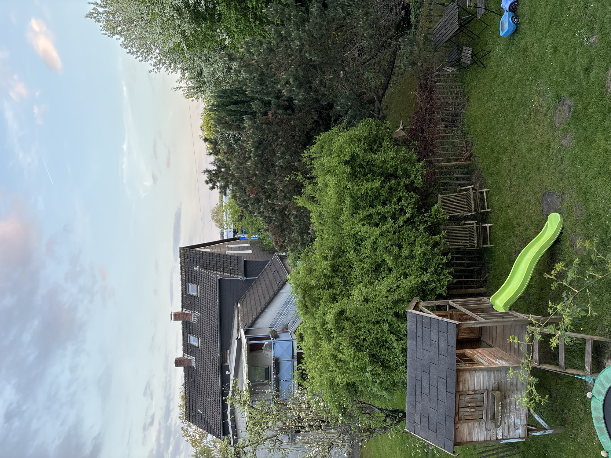 This elevated view captures a residential backyard scene, featuring a house on the left and a well-maintained garden space on the right, under a bright, partly cloudy sky.
The prominent subject on the left is a multi-story house with a dark, tiled gabled roof, featuring several brick chimneys and dormer windows. Below the main roofline, a glass-enclosed balcony or sunroom extends from the house, surrounded by lush green foliage and a tree with white blossoms in the foreground.
The right side of the image reveals a vibrant green lawn. A wooden playhouse with a dark shingled roof stands prominently in the lower right, equipped with a bright green slide. Partially visible at the very bottom right is a trampoline. Further into the yard, a small blue ride-on toy is visible on the grass, near a wooden outdoor dining set consisting of a table and several chairs. A series of wooden steps or terraced planters ascend a gentle slope, bordered by dense green bushes and various trees, including conifers and leafy varieties, creating a verdant backdrop.
The sky above is a soft light blue with scattered white and grey clouds, and a faint airplane contrail is visible. The lighting suggests it is daytime, possibly late afternoon, creating a serene and inviting atmosphere. No people are present, indicating a tranquil, domestic setting prepared for leisure and play. There is no visible text in the image.
