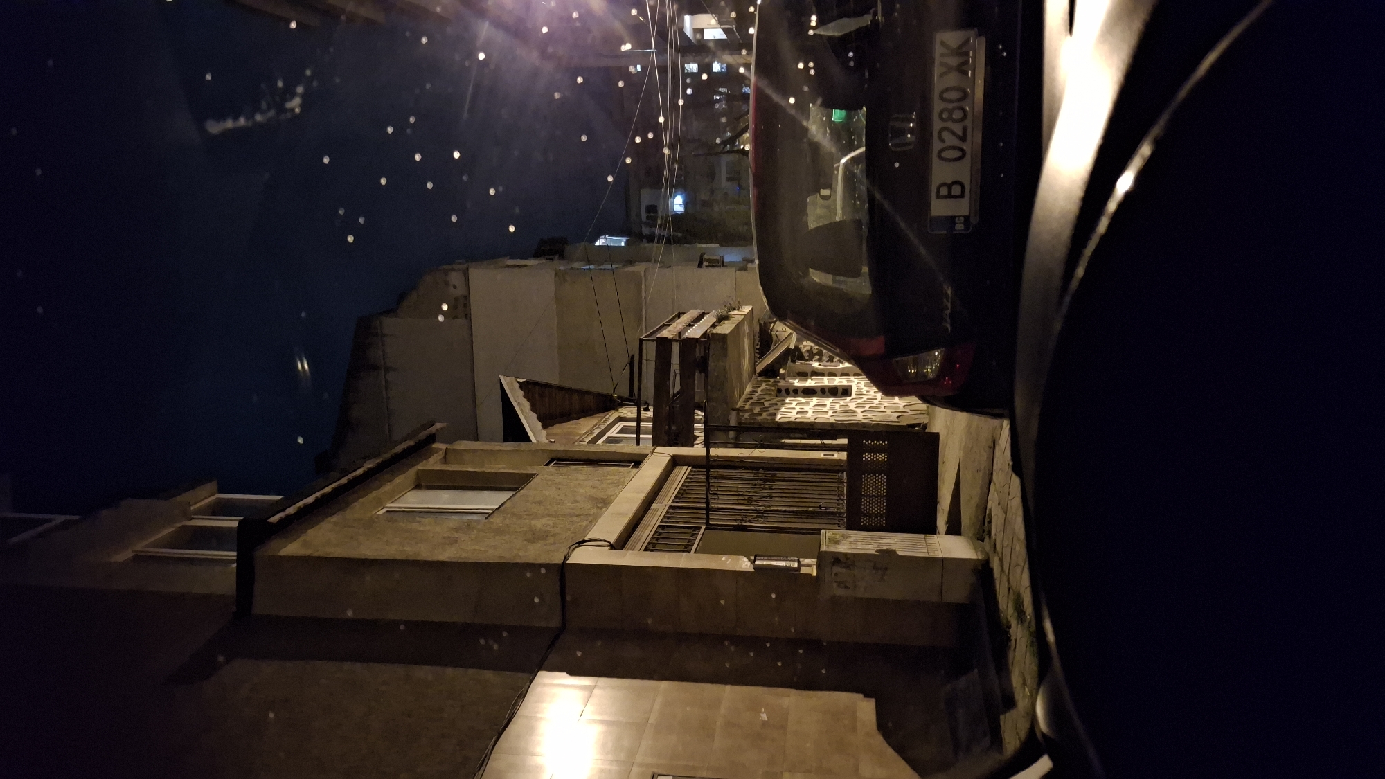 The image, taken at night, shows a view of buildings and a partially visible car. The buildings are multi-storied and appear to be made of concrete or stone, with visible windows and balconies. The area is lit by streetlights and possibly interior lights from the buildings, creating a contrasting play of light and shadow. The car is positioned near the edge of the frame, with its license plate "B 0280 XK" visible. The overall composition suggests a scene captured from an elevated vantage point, possibly a balcony or window. The specific location within the city of Varna, Bulgaria, cannot be determined without additional information or context.