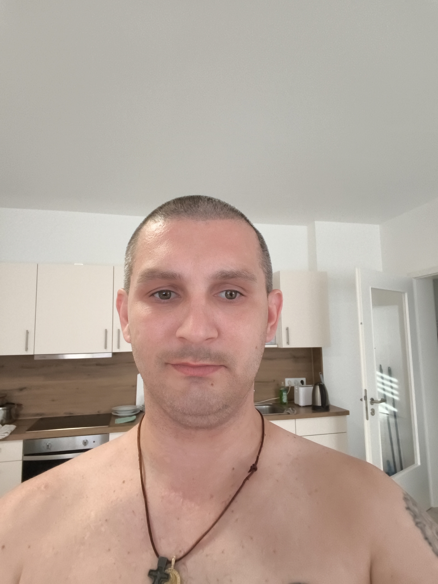 A man is shown from the chest up, looking directly at the camera. He has short, dark hair and a fair complexion. He is not wearing a shirt, but a brown cord with a pendant is visible around his neck. His shoulders and upper chest show some minor skin irregularities.
The background appears to be a domestic kitchen. Light-colored cabinets are mounted on the wall, with a wooden countertop below. A stovetop and oven are visible, as are kitchen appliances like a kettle and what appears to be a toaster. A doorway with a glass panel is visible to the right, leading to another room. The lighting suggests it is daytime.
The location is identified as Wolgast, Germany.