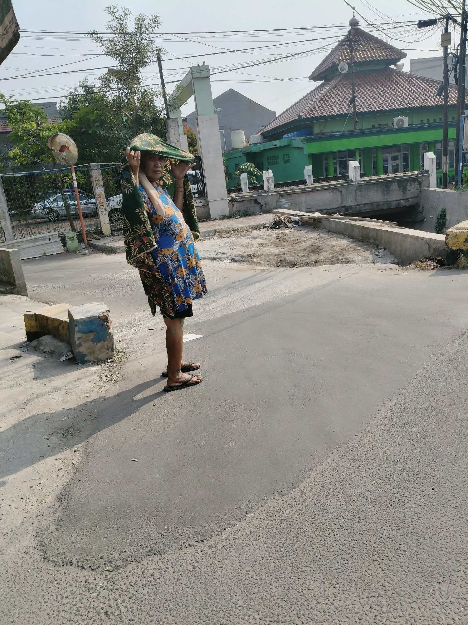 An adult, wearing a patterned blue dress and a green headscarf, stands on an asphalt road in South Tangerang, Indonesia. The person is holding the headscarf with both hands, partially obscuring their face. They are barefoot, wearing flip-flops. In the background, a green mosque with a tiled roof is visible, along with a bridge and trees. The scene appears to be outdoors in a residential or semi-urban area. The lighting suggests it is daytime.