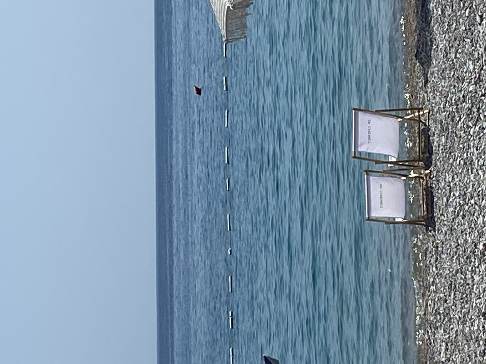 Two white canvas beach chairs with wooden frames are set up on a pebble beach facing the blue ocean. The chairs are positioned close together, and one has a visible logo that reads "M CHENEL". In the background, the calm ocean stretches to the horizon under a clear blue sky. A line of buoys floats on the water in the middle ground, and a small red triangular marker is visible further out. To the far right, the edge of a white and striped beach umbrella is visible. The scene appears to be a tranquil beach setting. The image was captured in Prijevor, Montenegro.